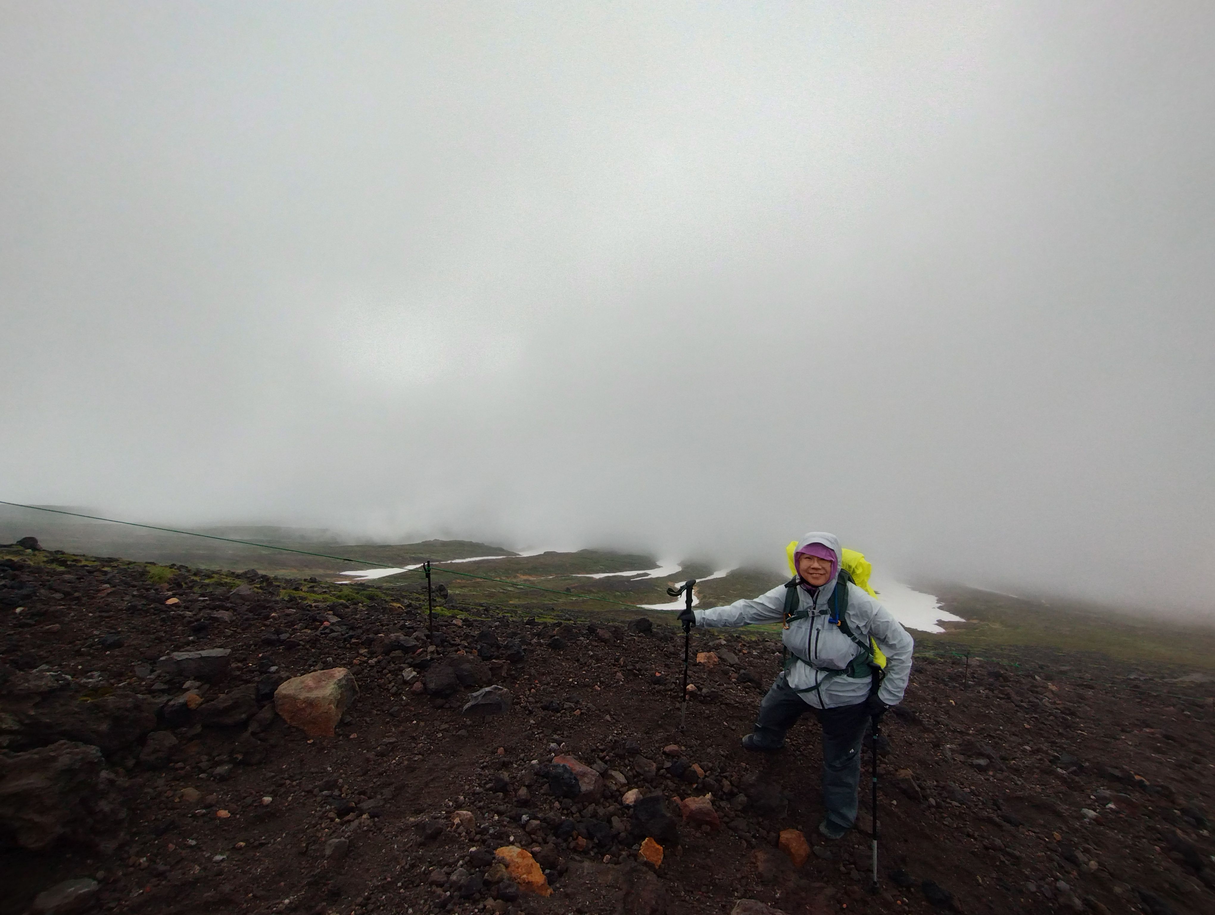 A hiker stands on a rocky mountain trail in rain gear on the Mt. Asahidake trail in Hokkaido. Snowfields are visible on the mountain behind them.