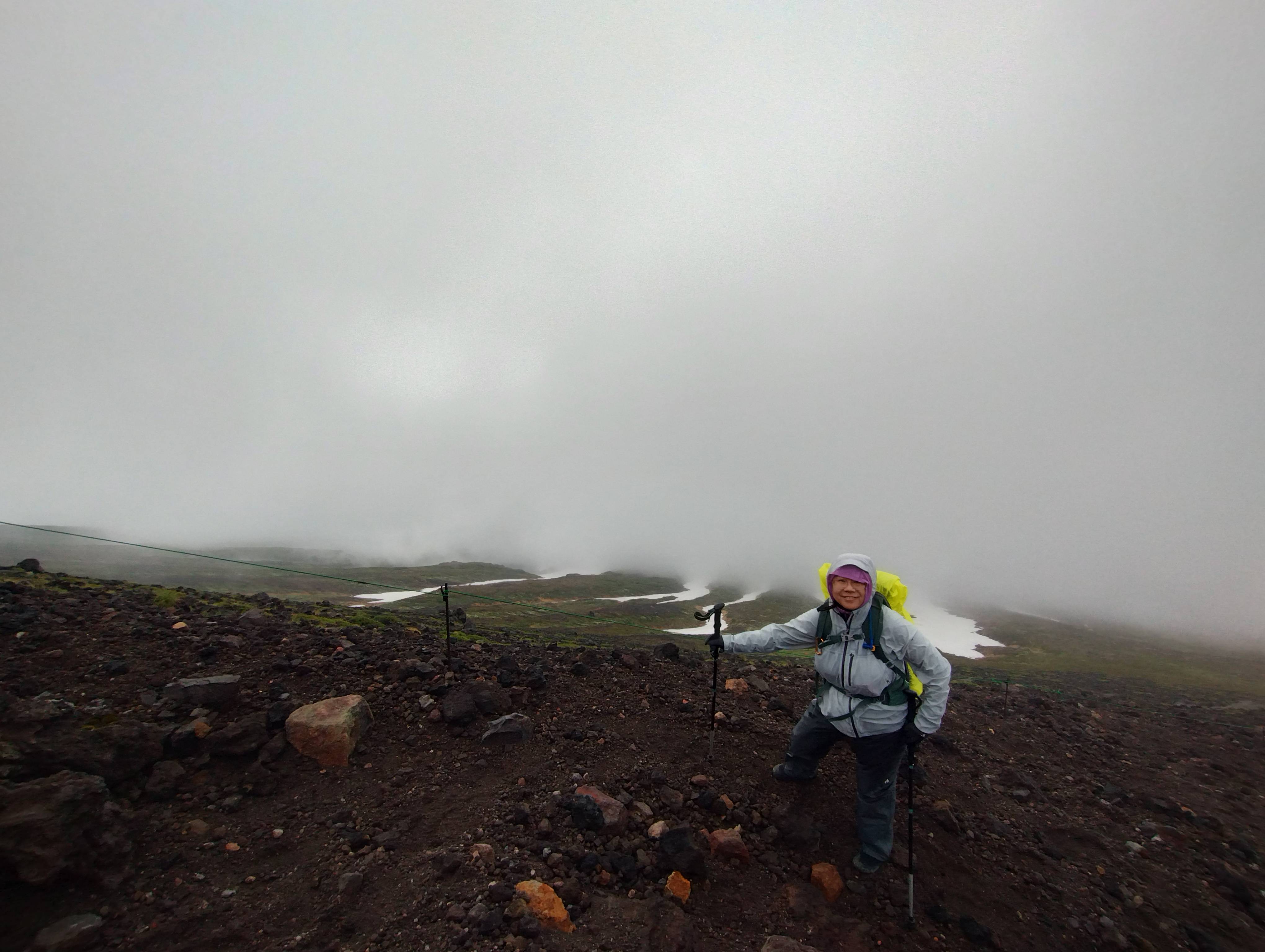 A hiker stands on a rocky mountain trail in rain gear on the Mt. Asahidake trail in Hokkaido. Snowfields are visible on the mountain behind them.