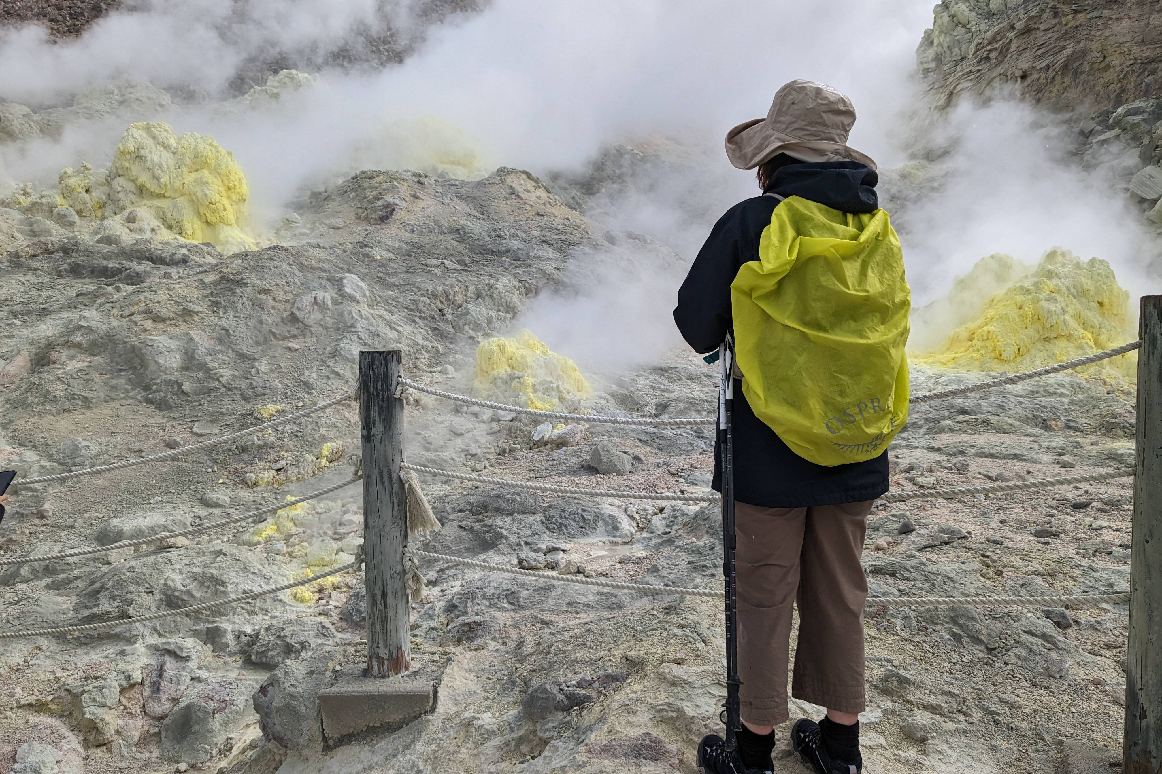 A tourist with a yellow pack cover stands in front of steaming volcanic vents that are coated in yellow sulpher