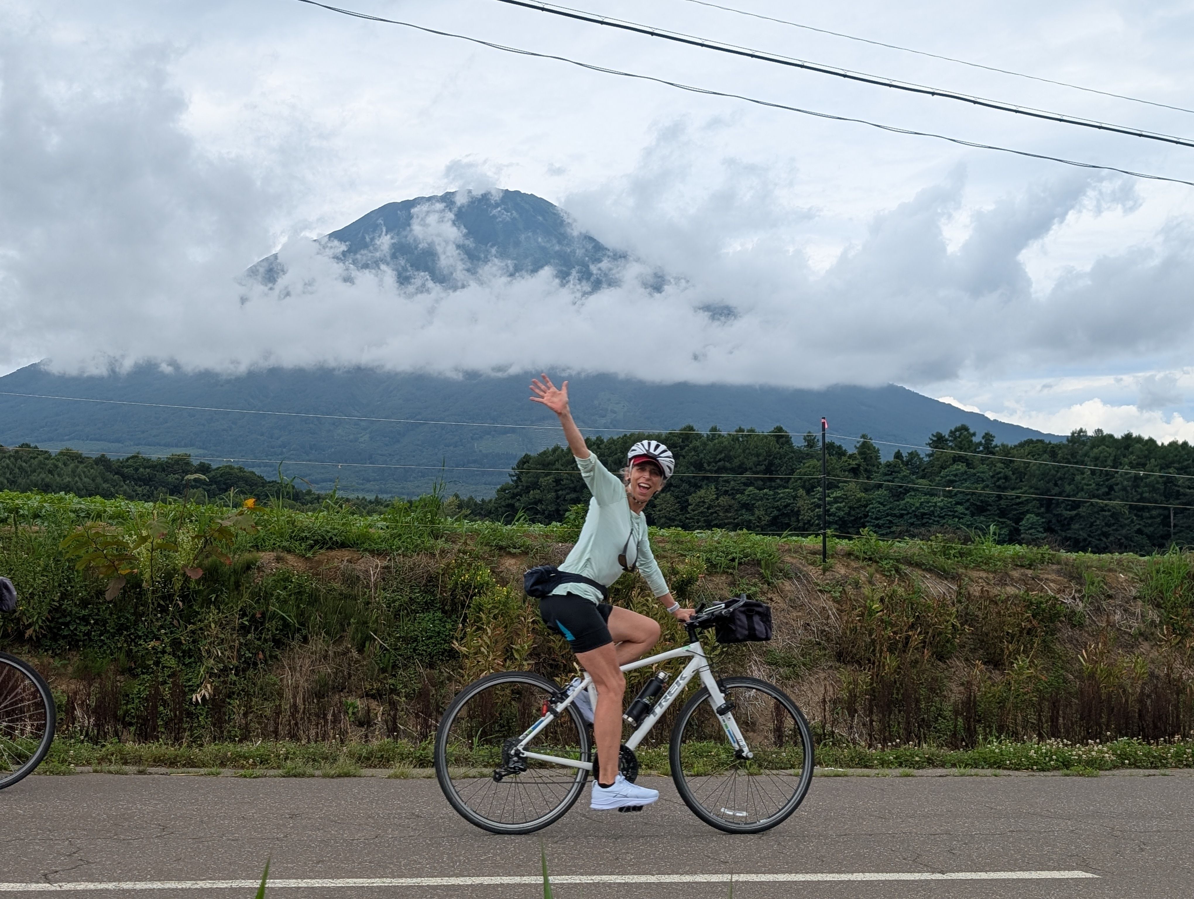 A woman on a bike waves at the camera as she passes Mt. Yotei in Hokkaido. The mountain is framed by thin clouds.