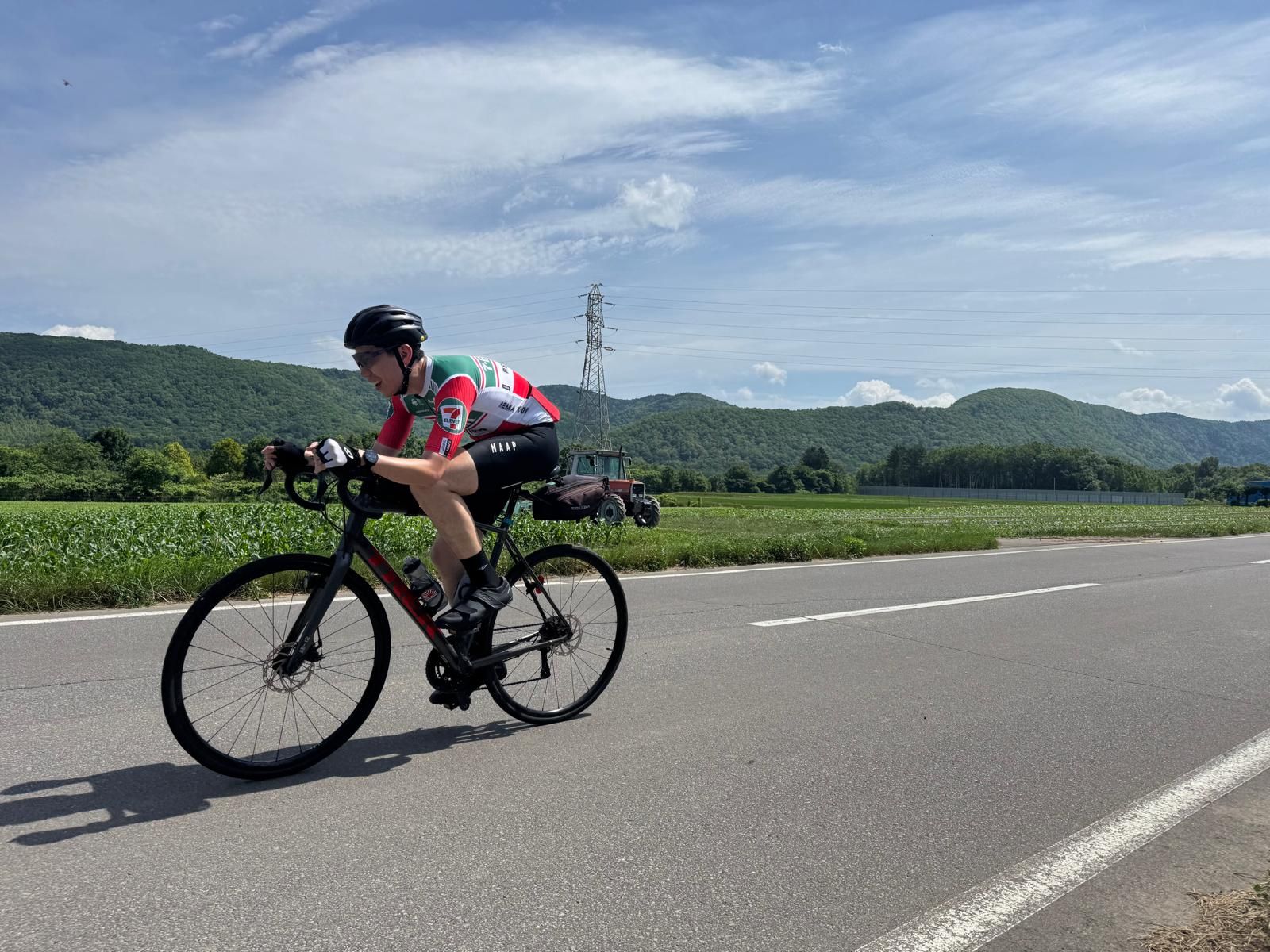 A cyclist cycles along a main road in rural Asahikawa. Flat farmland is visible around him, along with distant mountains.