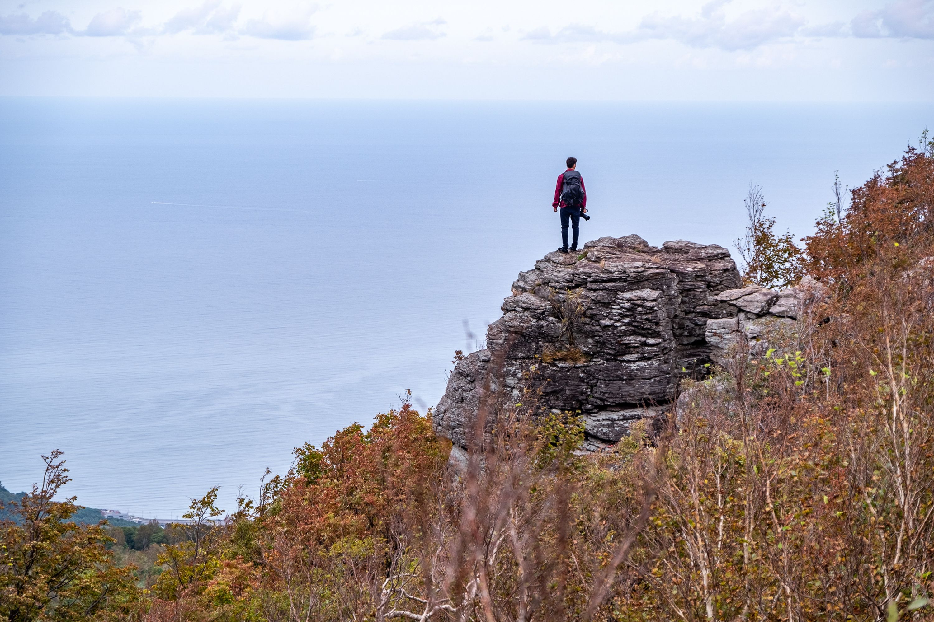 A lone hiker stands on a rocky summit on Mt. Shioya-maruyama, looking out over the vast Sea of Japan. Autumn foliage frames the foreground under an overcast sky.