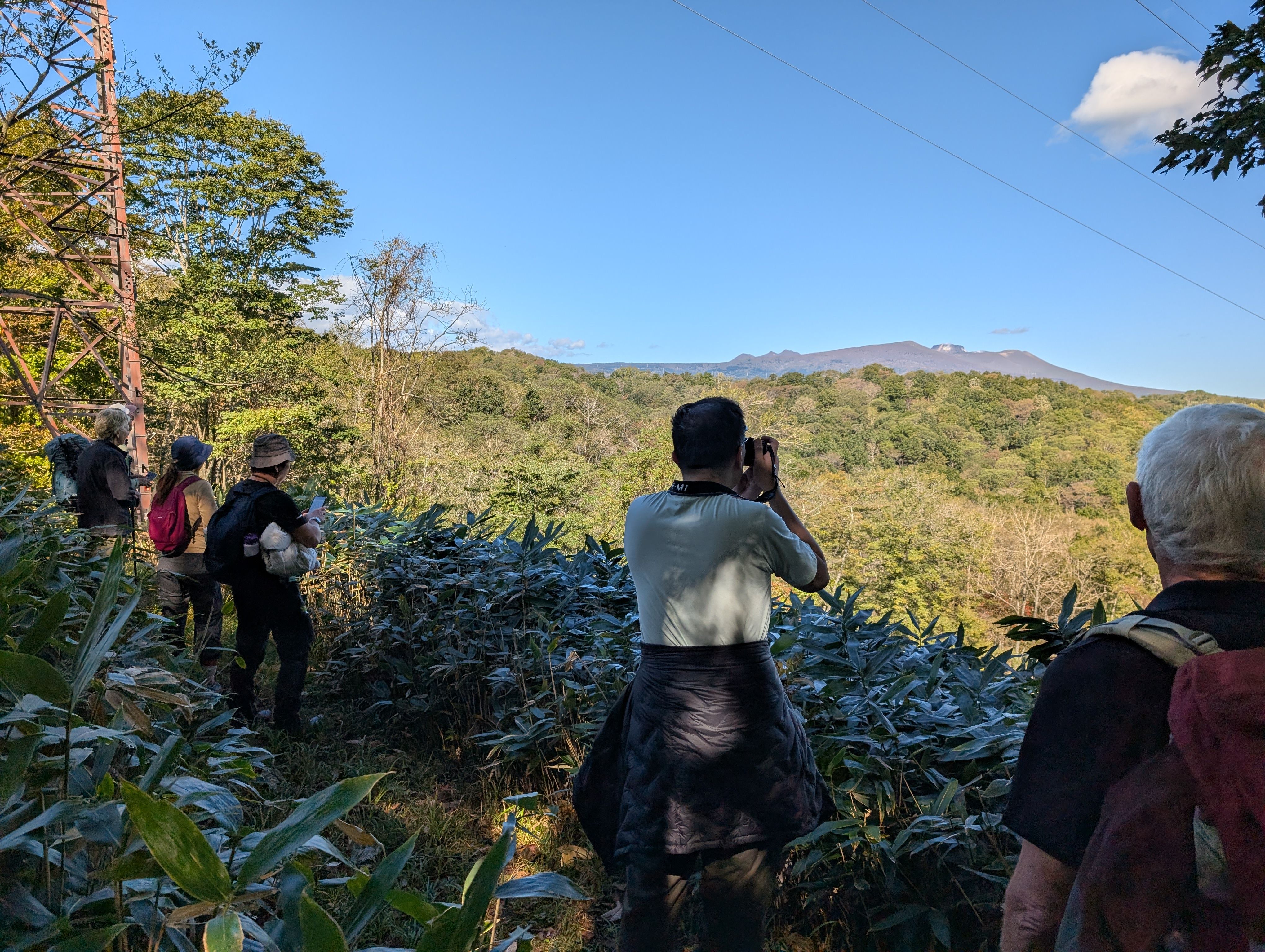 A group of hikers admire the view of Mt. Tarumae, visible from the highest point of the nature trail at Lake Poroto, Hokkaido. It is a sunny day and the mountain is clearly visible. Some are taking photographs on cameras and smartphones.