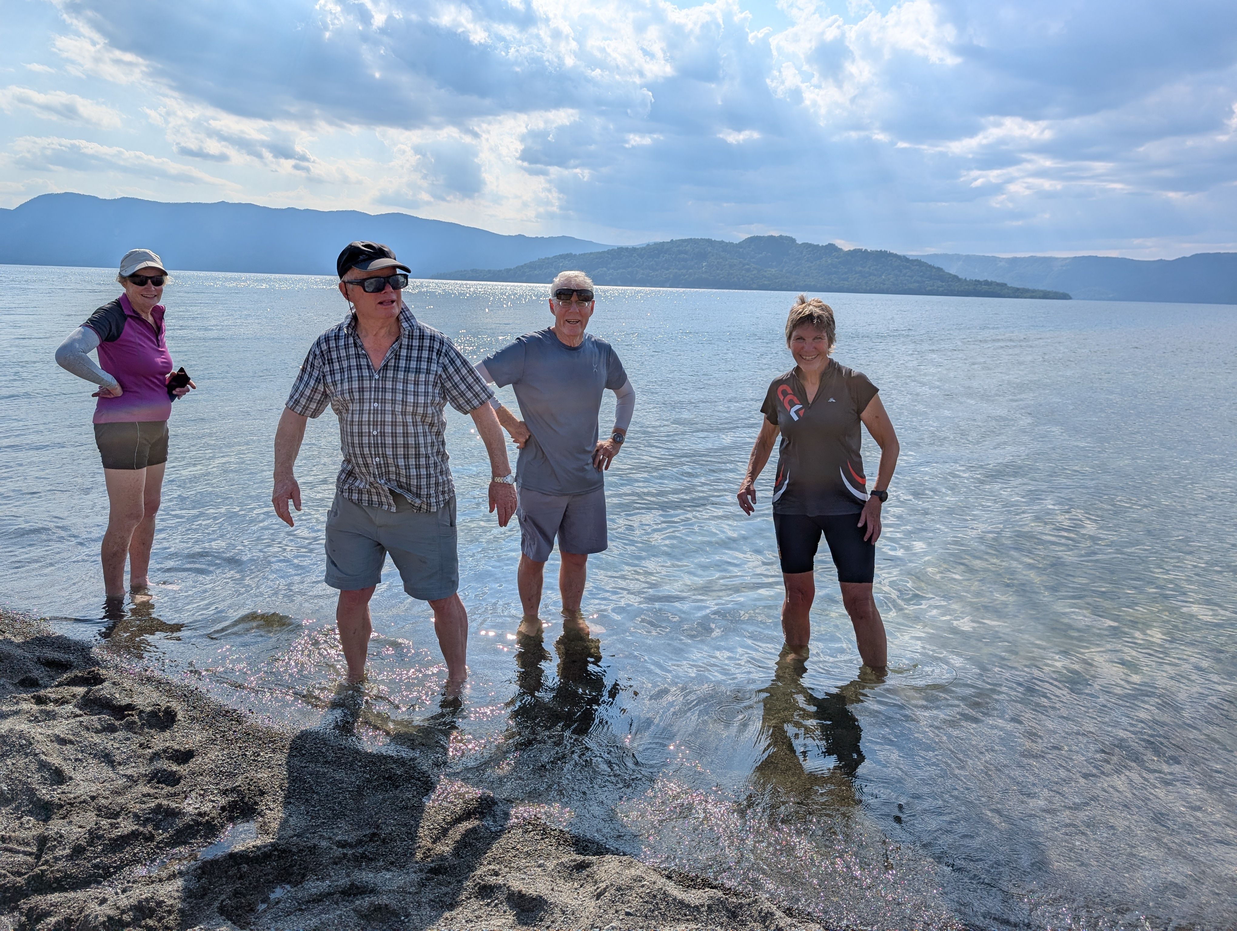 Four people stand on the shore of Lake Kussharo in Hokkaido. They are burying their feet in the sand, which is heated by underground hot springs.