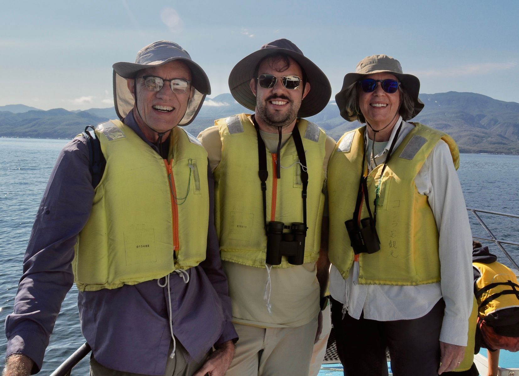 Three smiling guests wearing sun hats, sunglasses, and yellow life jackets pose together on a boat deck during a nature cruise on the Nemuro Strait. Two have binoculars around their necks. Calm blue waters and distant mountains are visible in the background under a bright, sunny sky.