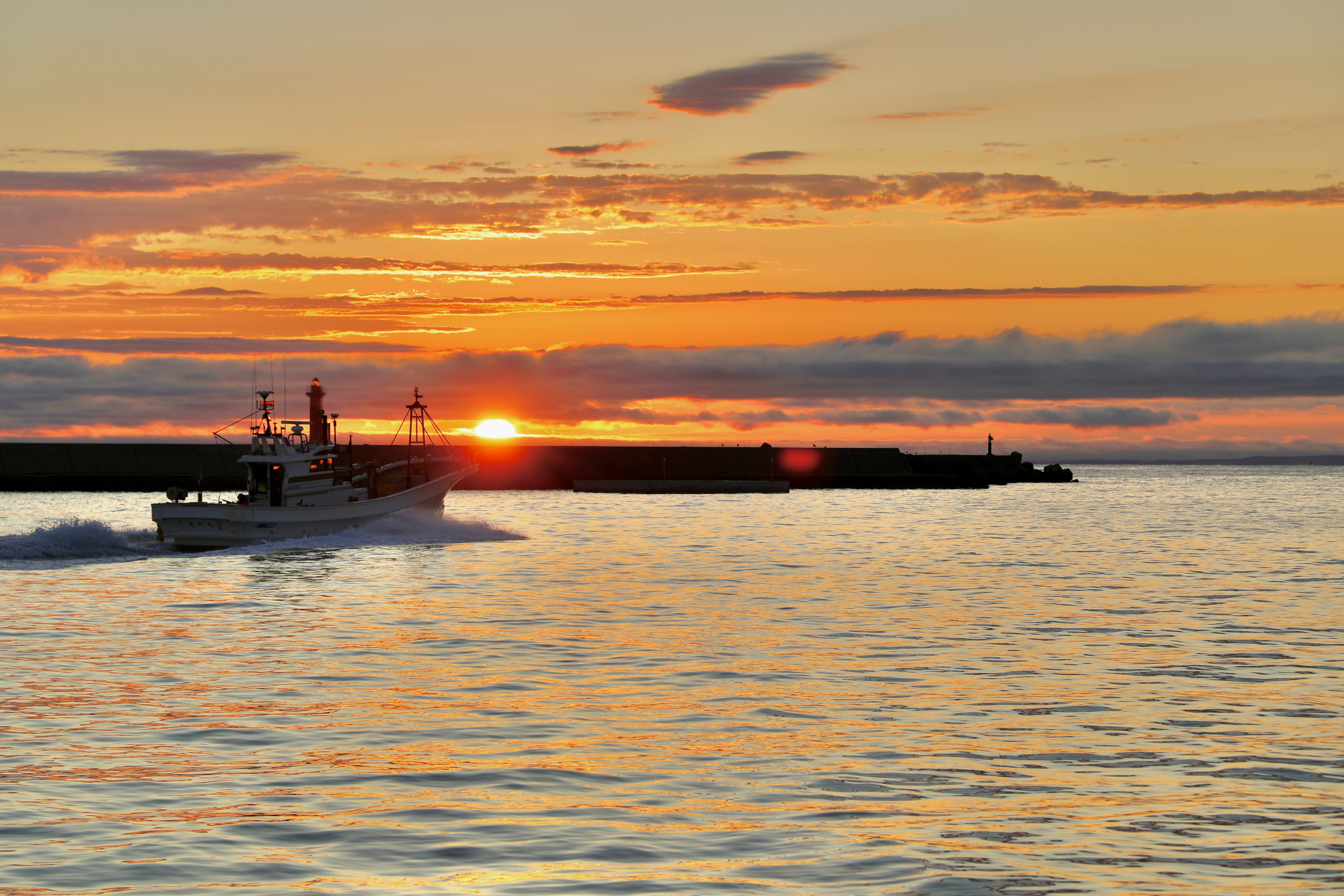 A photograph of a fishing boat leaving a harbour at sunset, heading out to sea. It is a very beautiful sunset and the sky is turning fiery colours. Photo credit: Samir Patel