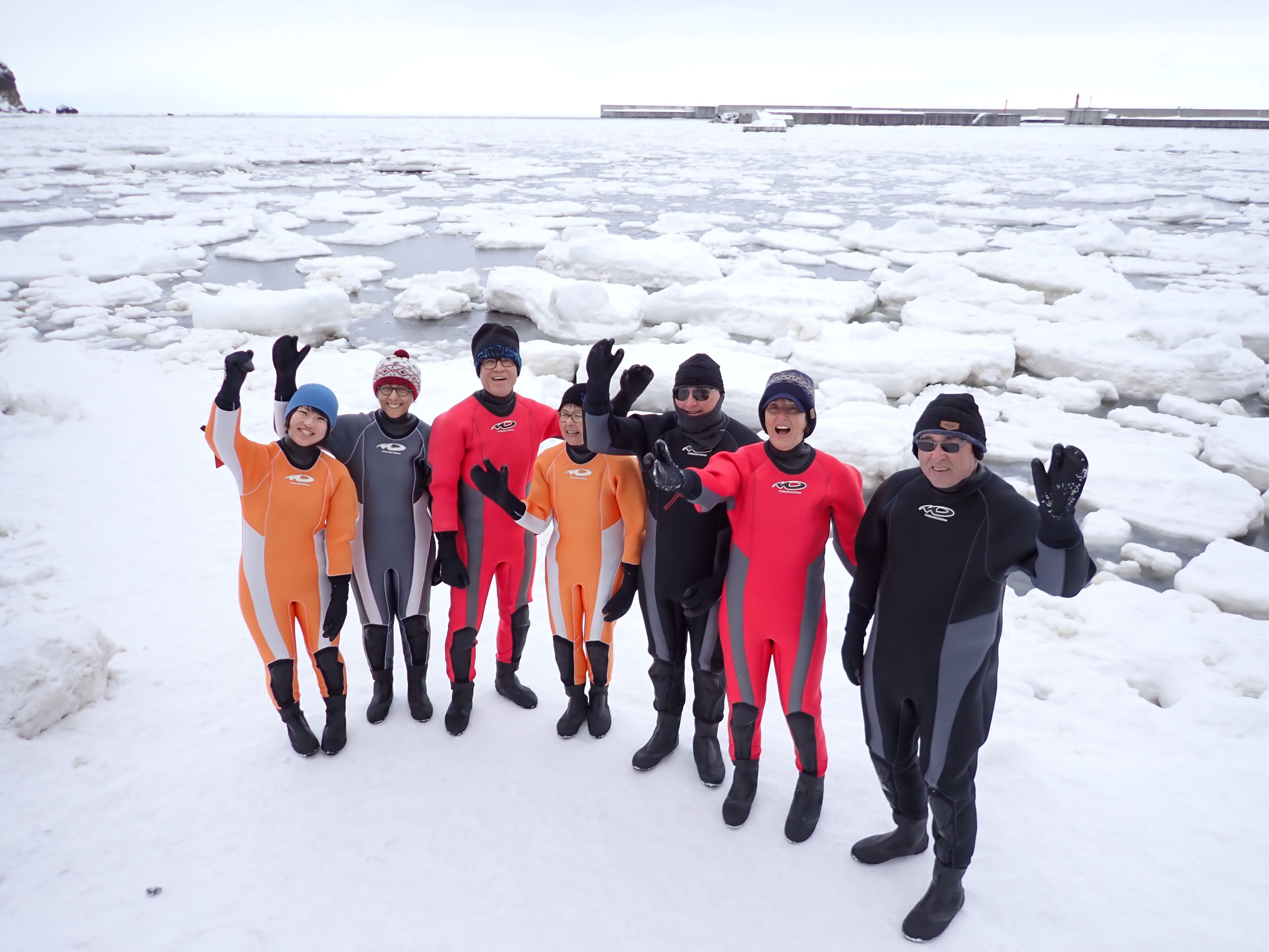 A group of people in drysuits stand at the edge of the ocean and smile and wave up at the camera. Behind them in the harbour are many floes of drift ice as far as the eye can see.