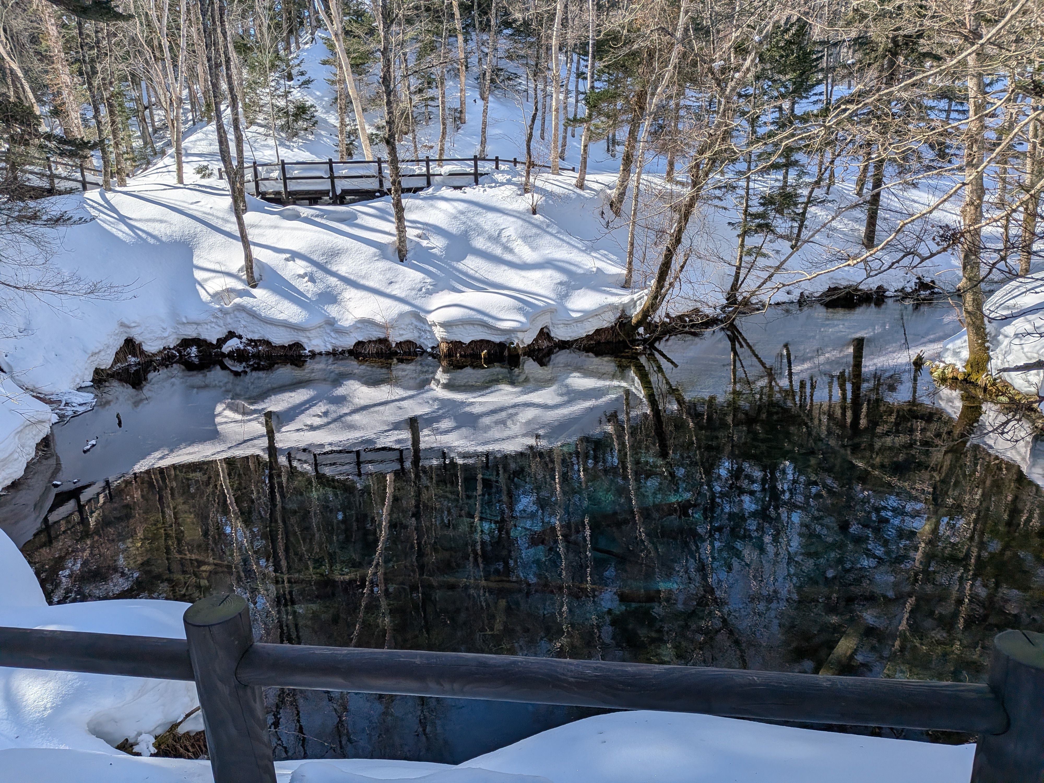 A photograph of Kaminokoike Pond in Hokkaido. The water is very clear and is a bright blue colour. Snow is on the banks around the pond.