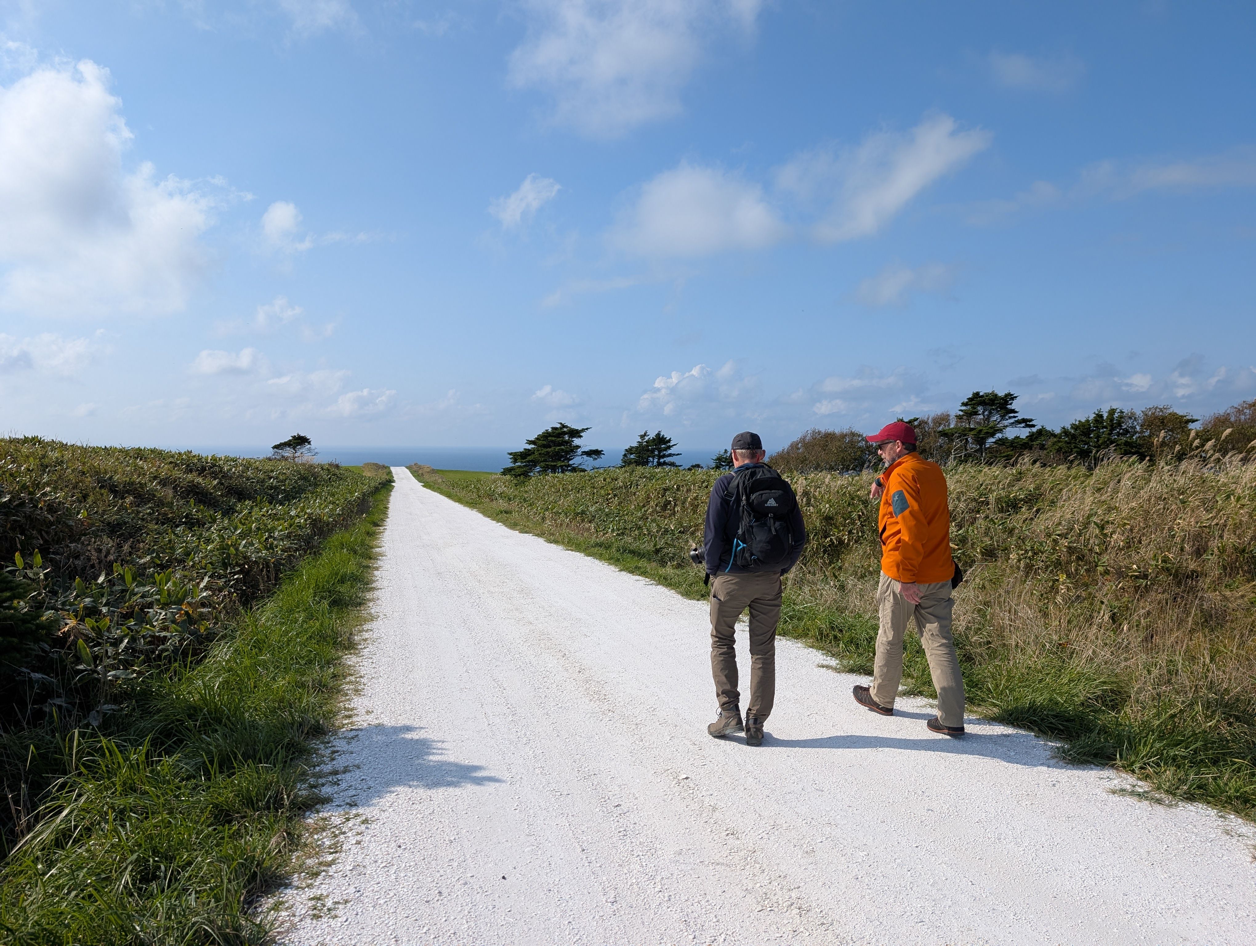 Two walkers walk along the White Shell Path (a small road made from crushed seashells) in Wakkanai, Hokkaido. The path stretches away to the horizon, where the ocean is visible. It is a sunny day.