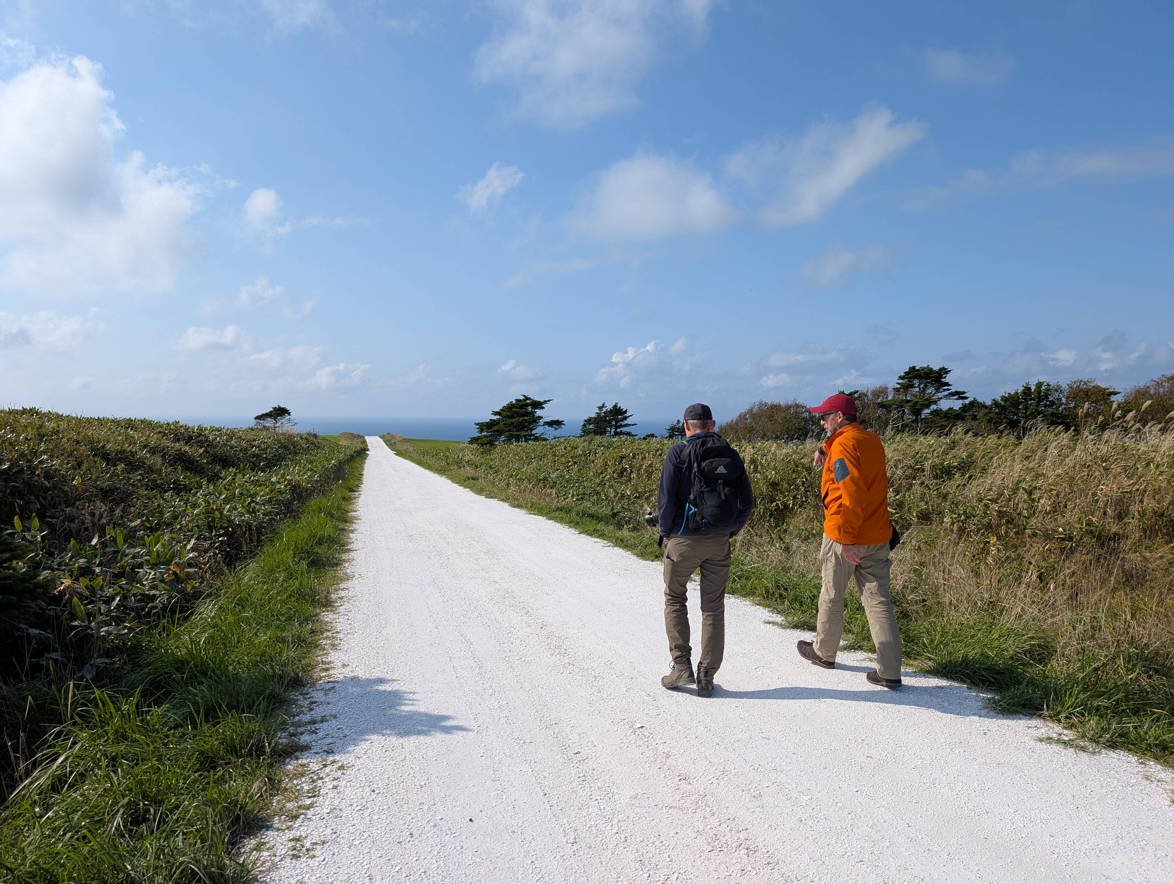 Two walkers walk along the White Shell Path (a small road made from crushed seashells) in Wakkanai, Hokkaido. The path stretches away to the horizon, where the ocean is visible. It is a sunny day.