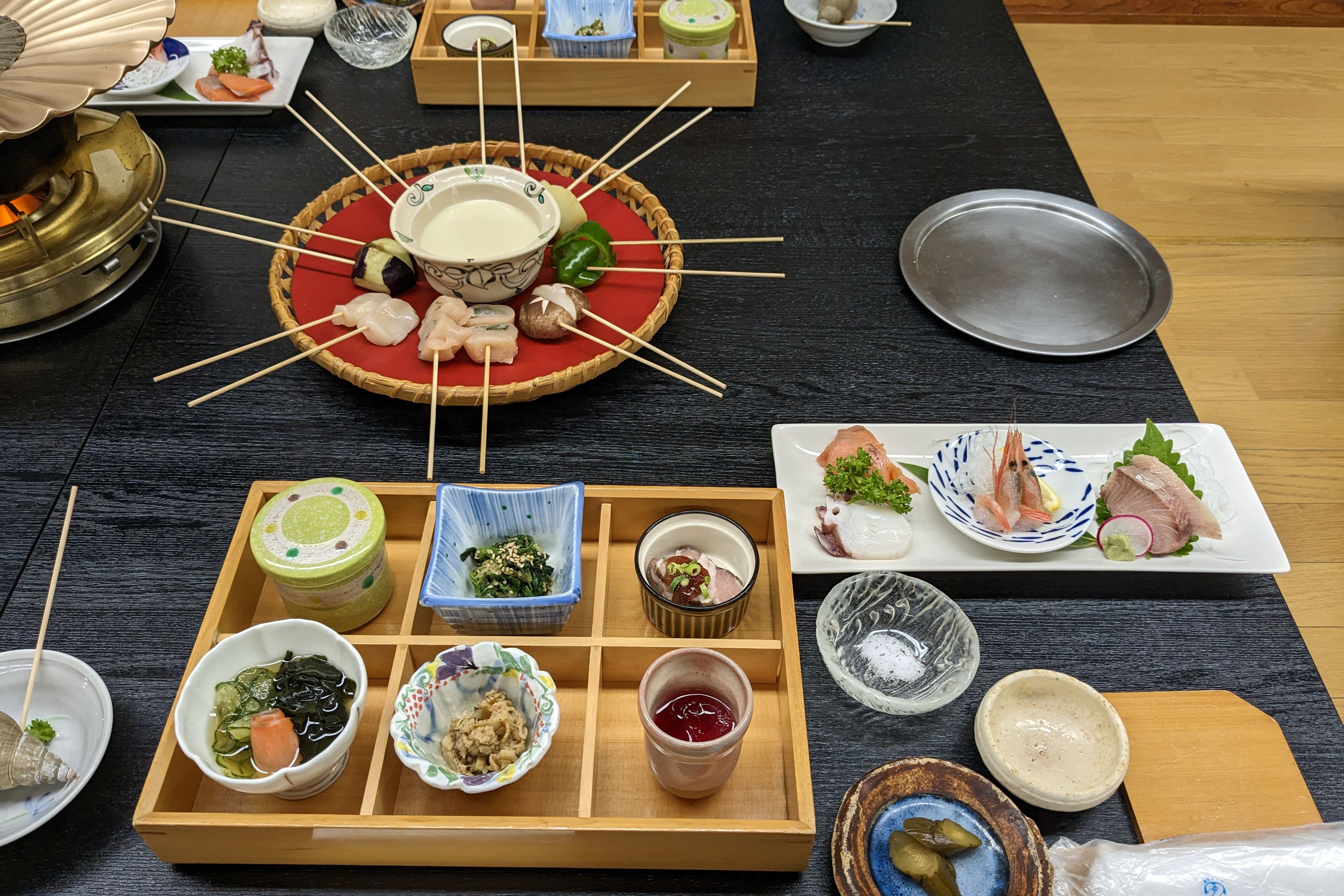 A feast is laid out on the table including meat and seafood on skewers, sashimi and small bowls.