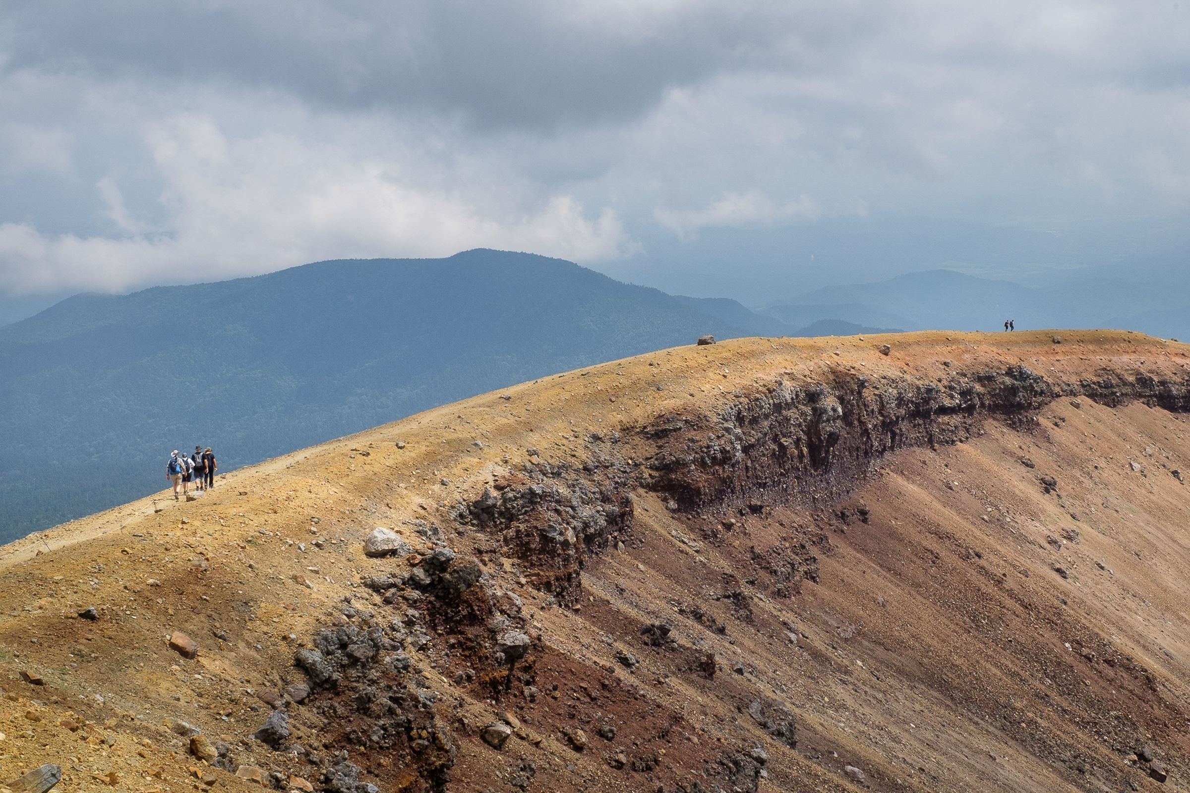 A group of hikers walking along the ridge of Mt Meakan are dwarfed by the rugged volcanic scenery. The sky is moody with large clouds. In the background a smaller peak sits in shade.