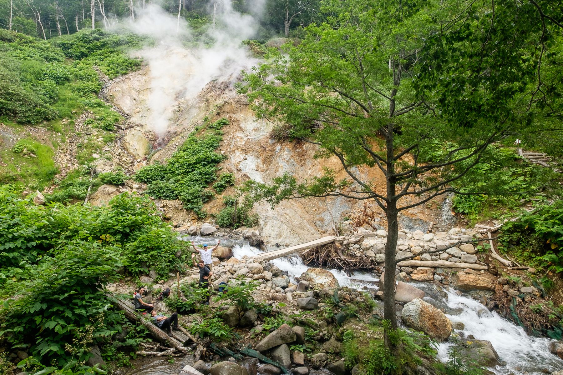 A group of hikers pose by a river, with a steam rising from a volcanic vent in the background