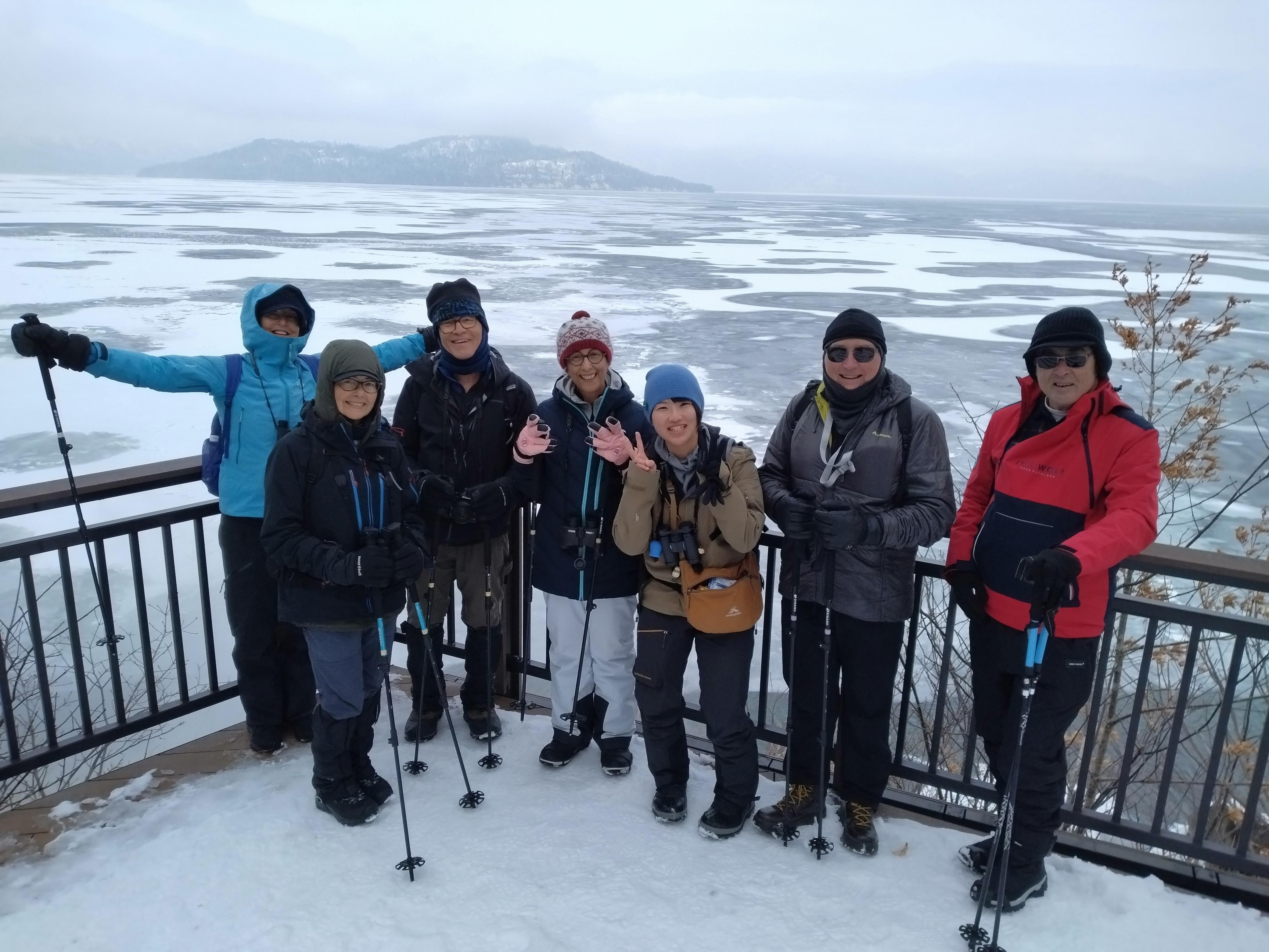 A group of people in winter gear smile and wave at the camera in front of a view of a frozen Lake Kussharo.