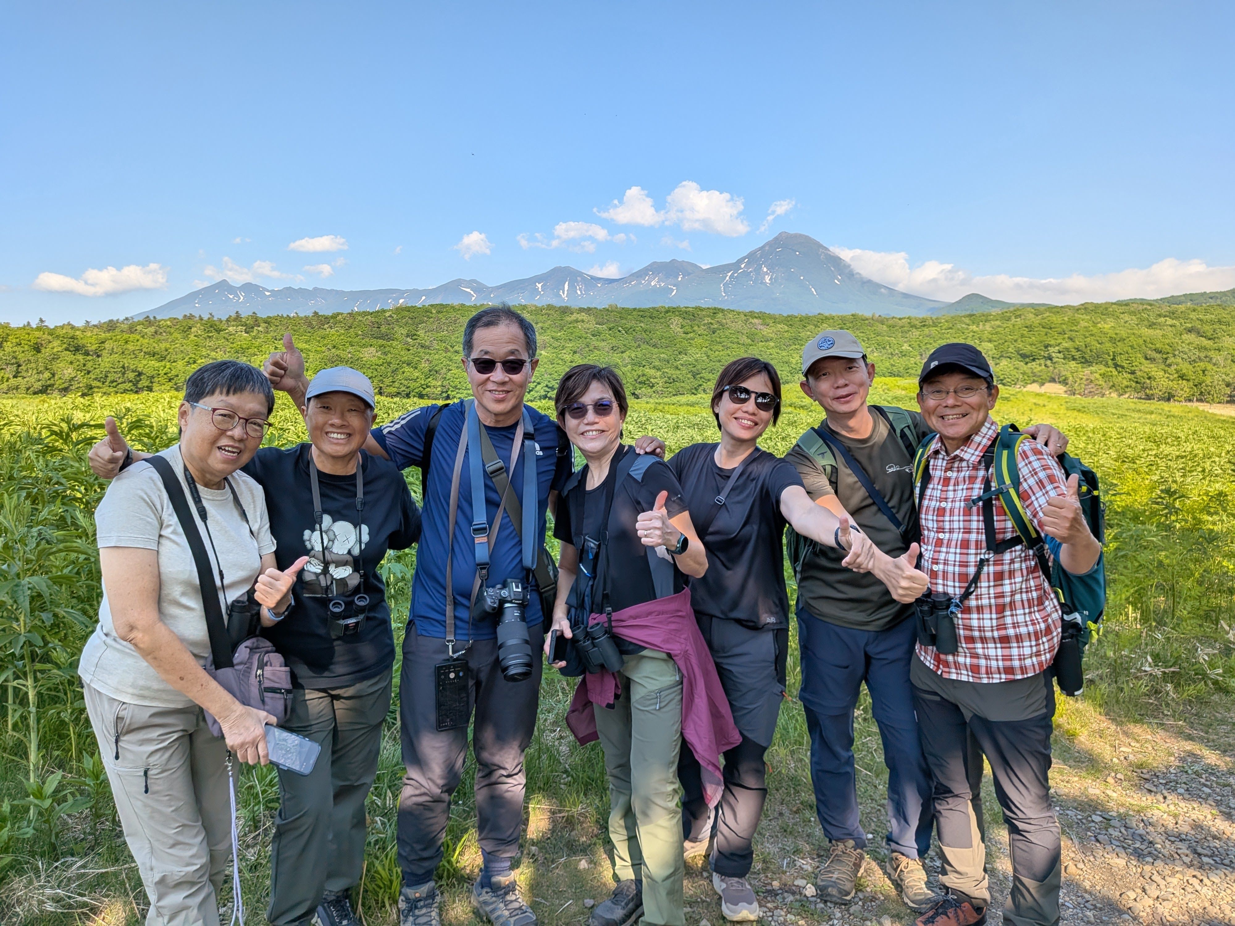 A group of people smile and give a thumbs-up at the camera. Behind them is a forest and the mountains of Shiretoko Peninsula, Hokkaido. It is a very sunny day and blue skies are visible beyond the mountains.