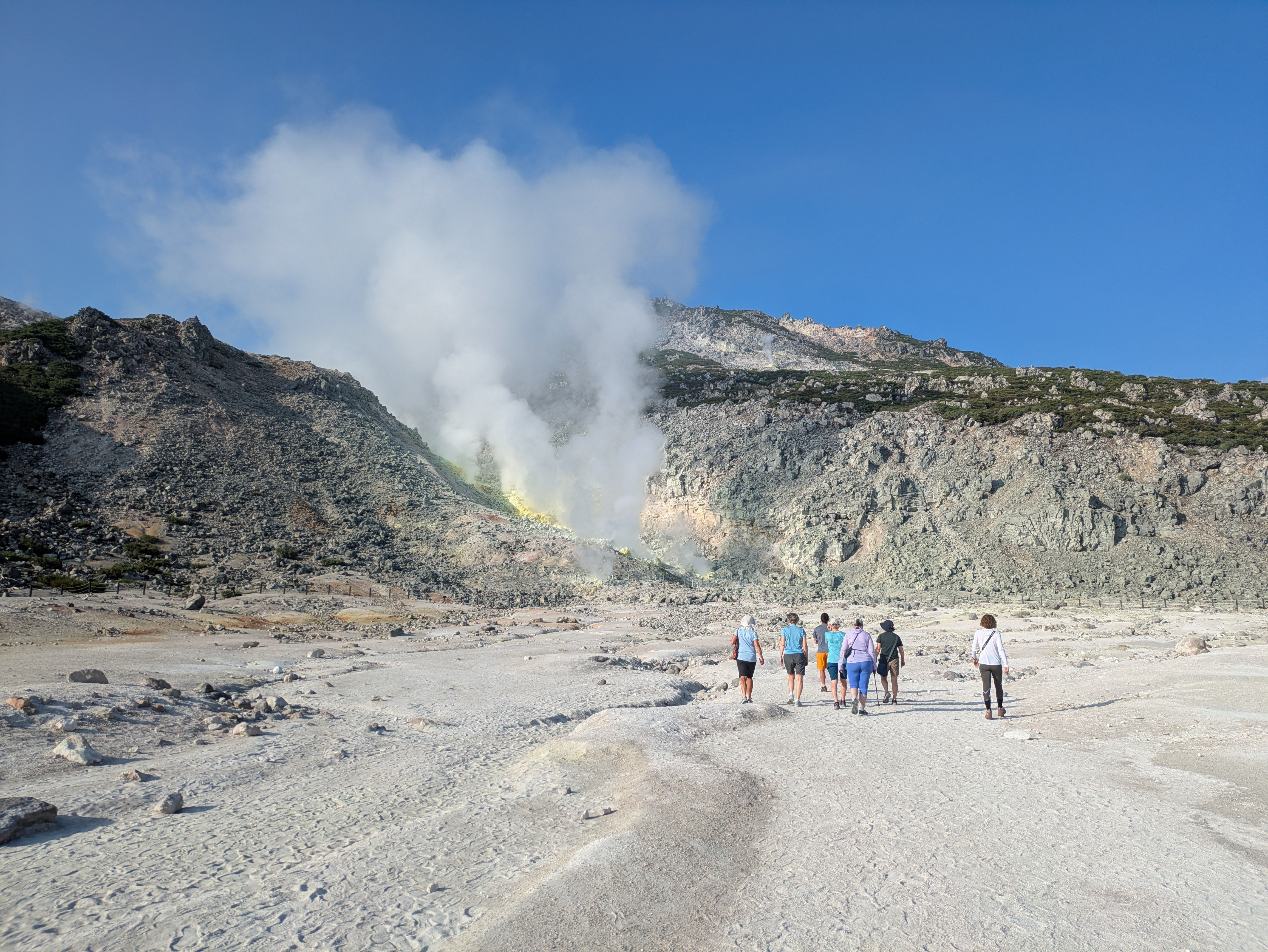 A group of hikers walk towards sulphuric fumaroles at Mt. Io, Hokkaido. It's a beautifully sunny day with no clouds in the sky. The neon yellow of sulphur is visible from the fumarole vents.