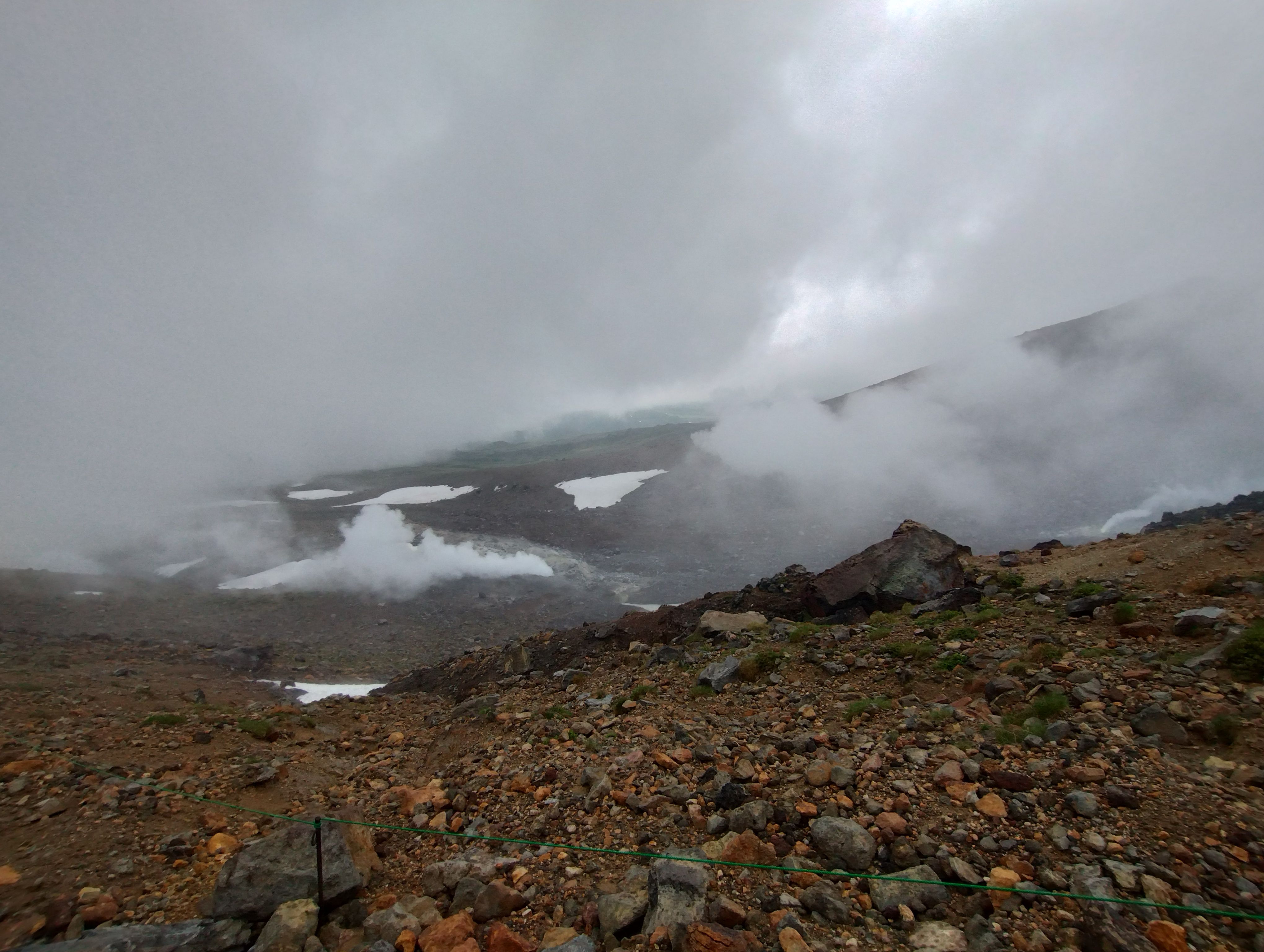 A photograph of Mt. Asahidake's fumaroles on a cloudy day. Steam rises from a volcanic crater on the mountain, snowfields in the background.