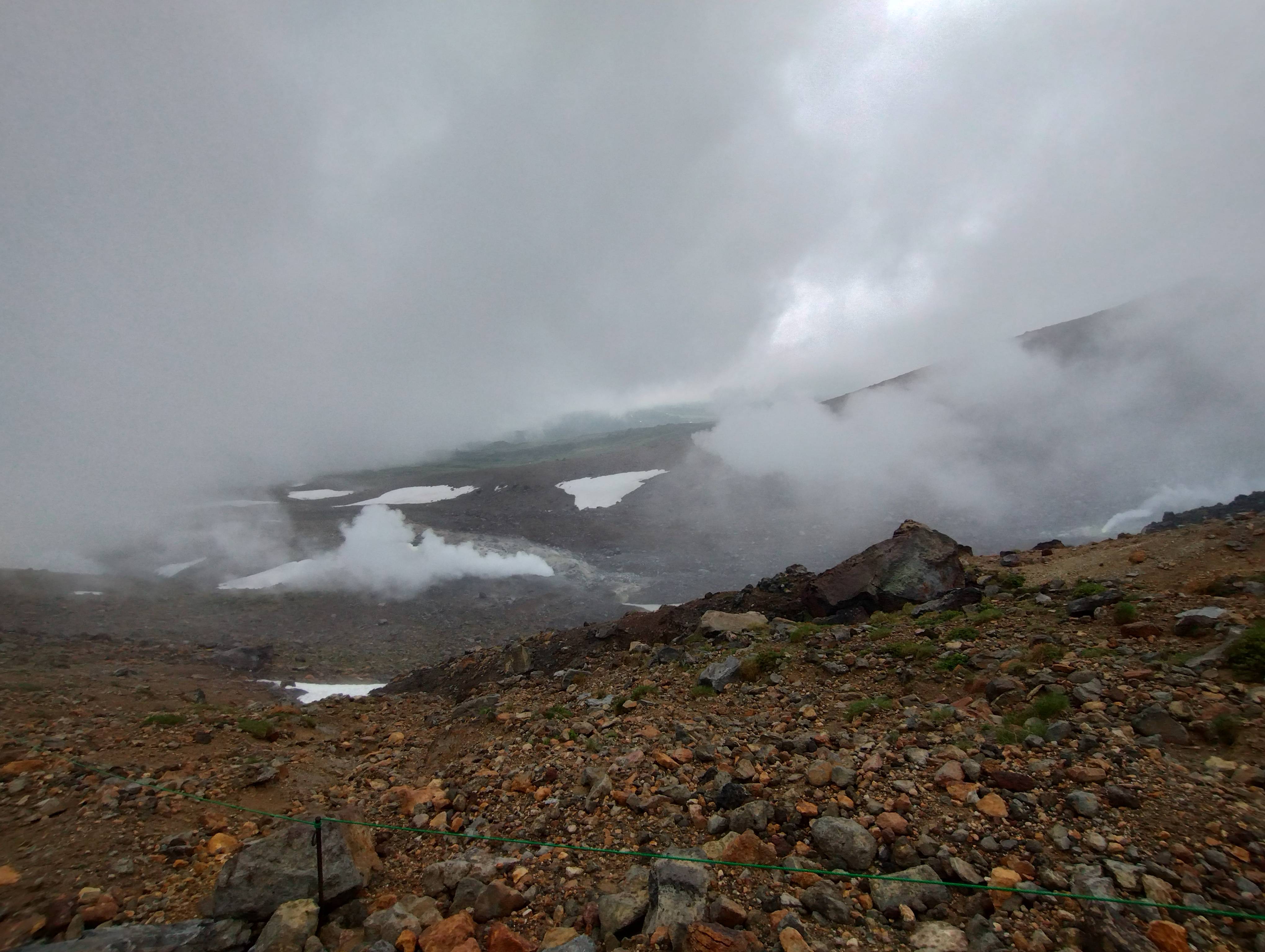 A photograph of Mt. Asahidake's fumaroles on a cloudy day. Steam rises from a volcanic crater on the mountain, snowfields in the background.
