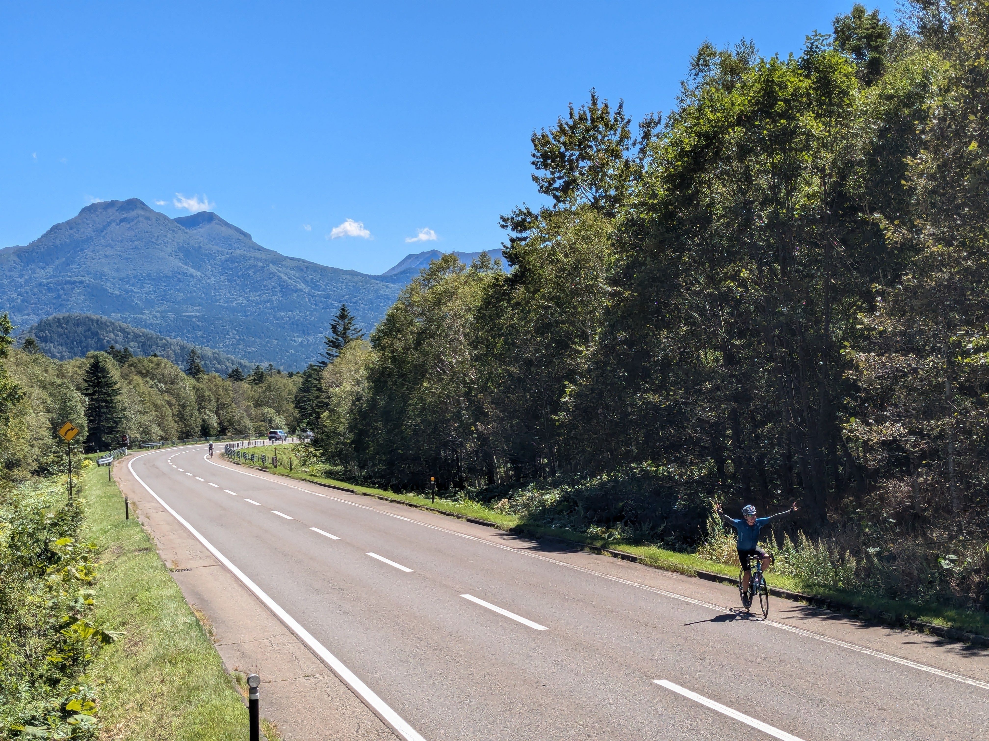 A cyclist rides along a road in Daisetsuzan National Park, Hokkaido. It is a sunny day and the peaks of mountains are visible in the background.