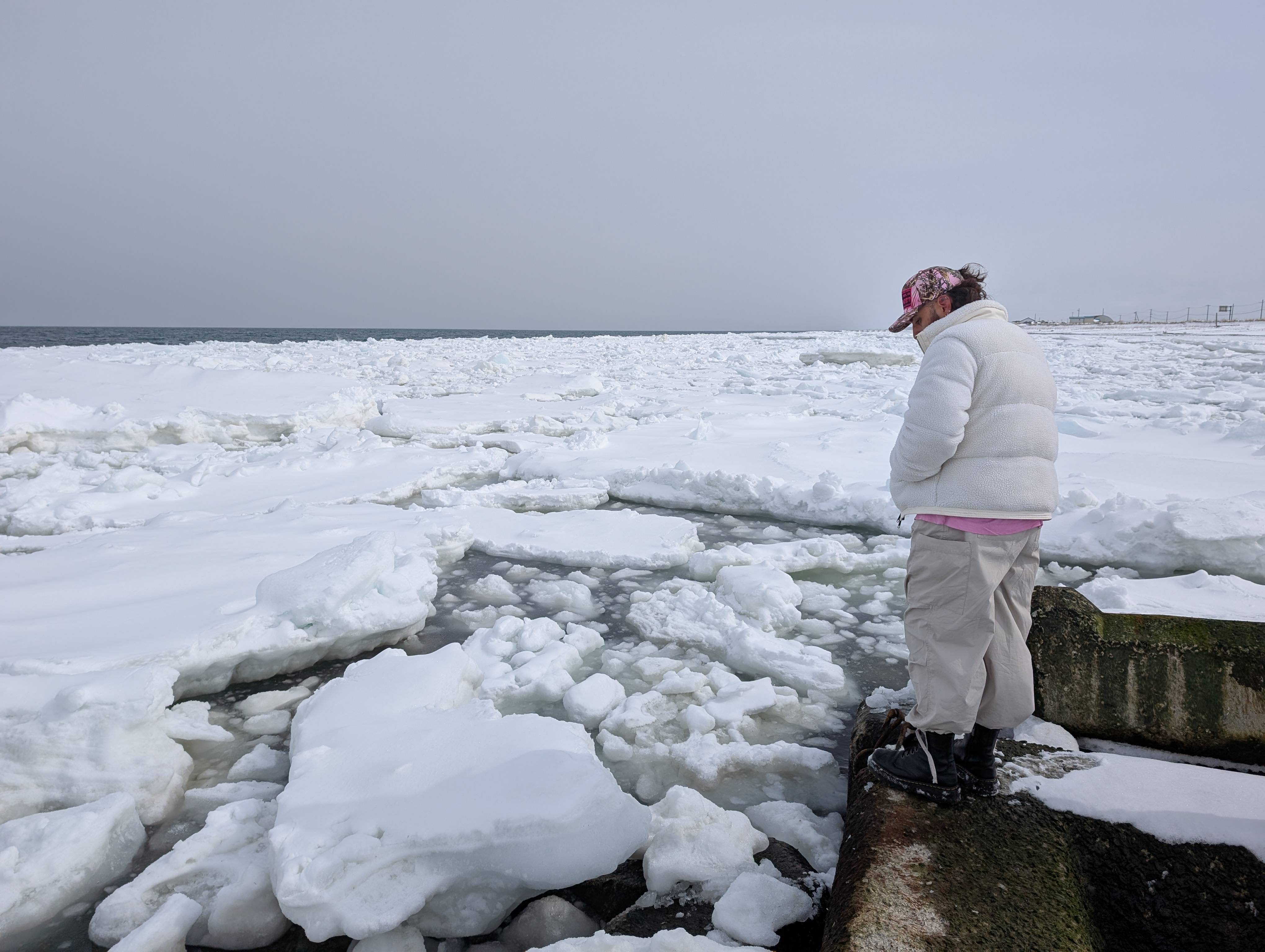 A man dressed in winter clothing stands at the edge of a sea wall, staring down at large pieces of drift ice floating in the sea below him.