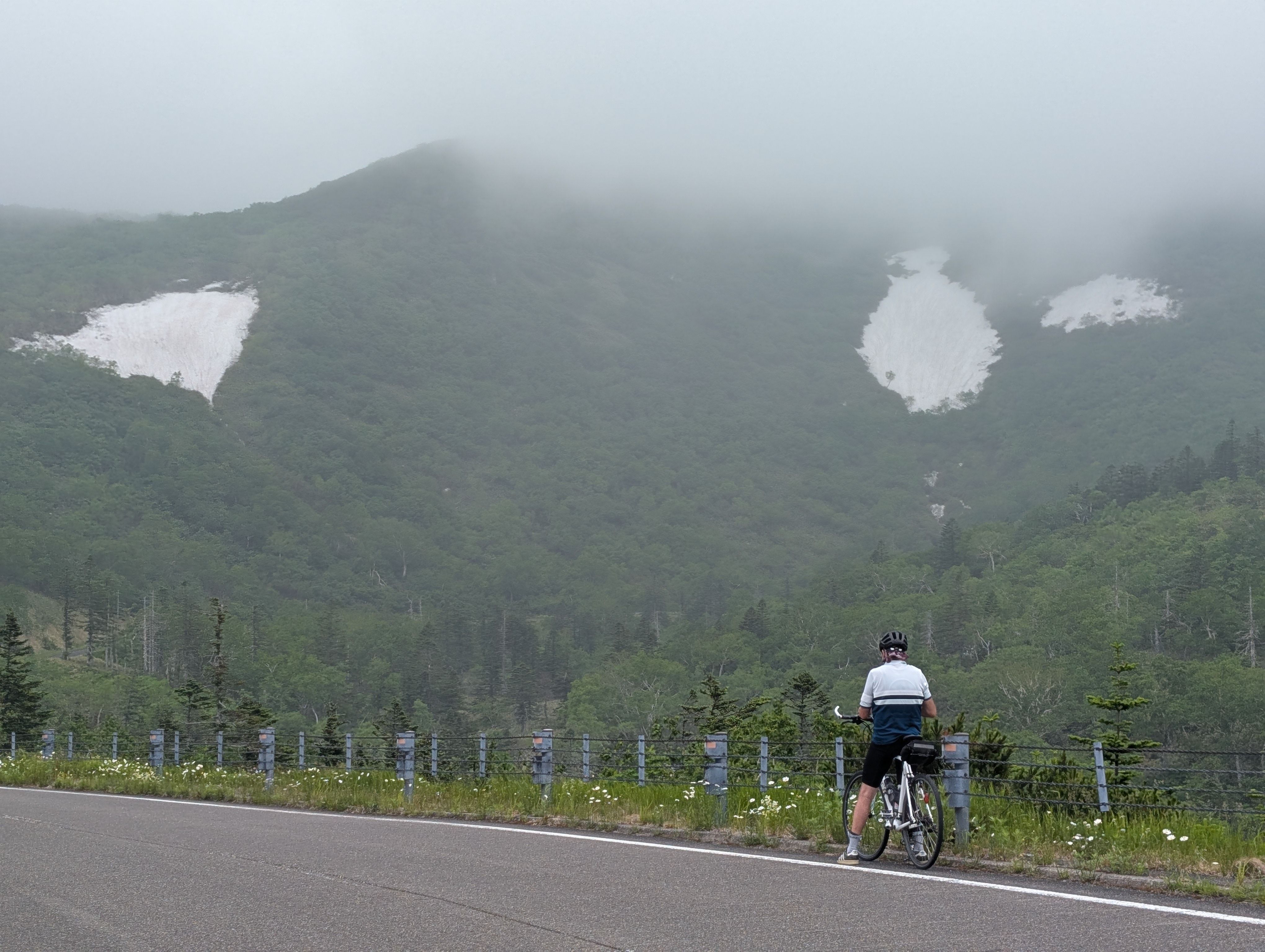 A man on a bike stops to take a look at mountains in the Shiretoko Pass, on which snow patches linger. It is a misty day and the tops of the mountains are obscured.