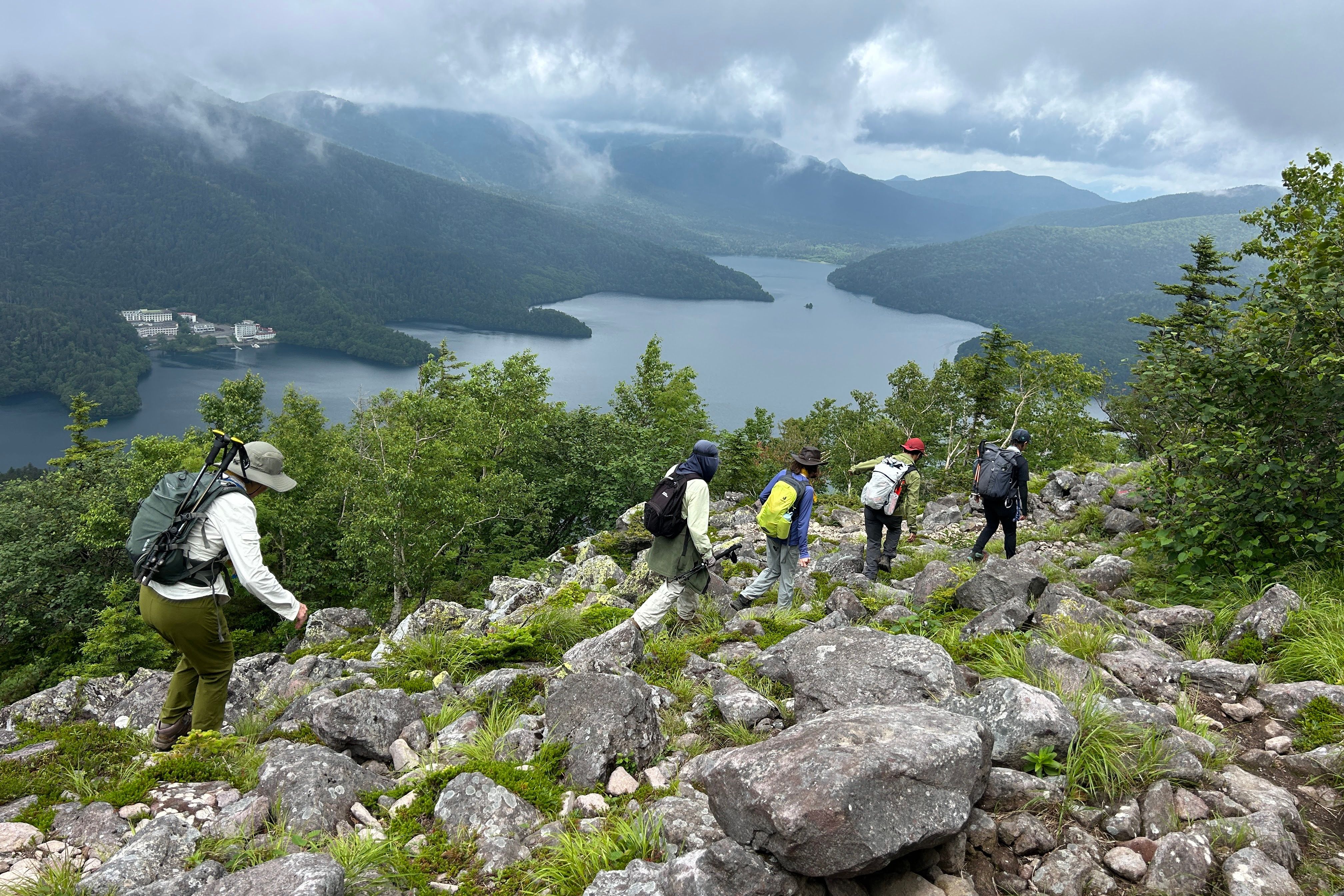 A group of hikers descend the rocky summit of Mt. Hakuunzan in Hokkaido. Below them, Lake Shikaribetsu is visible in a misty mountain valley.