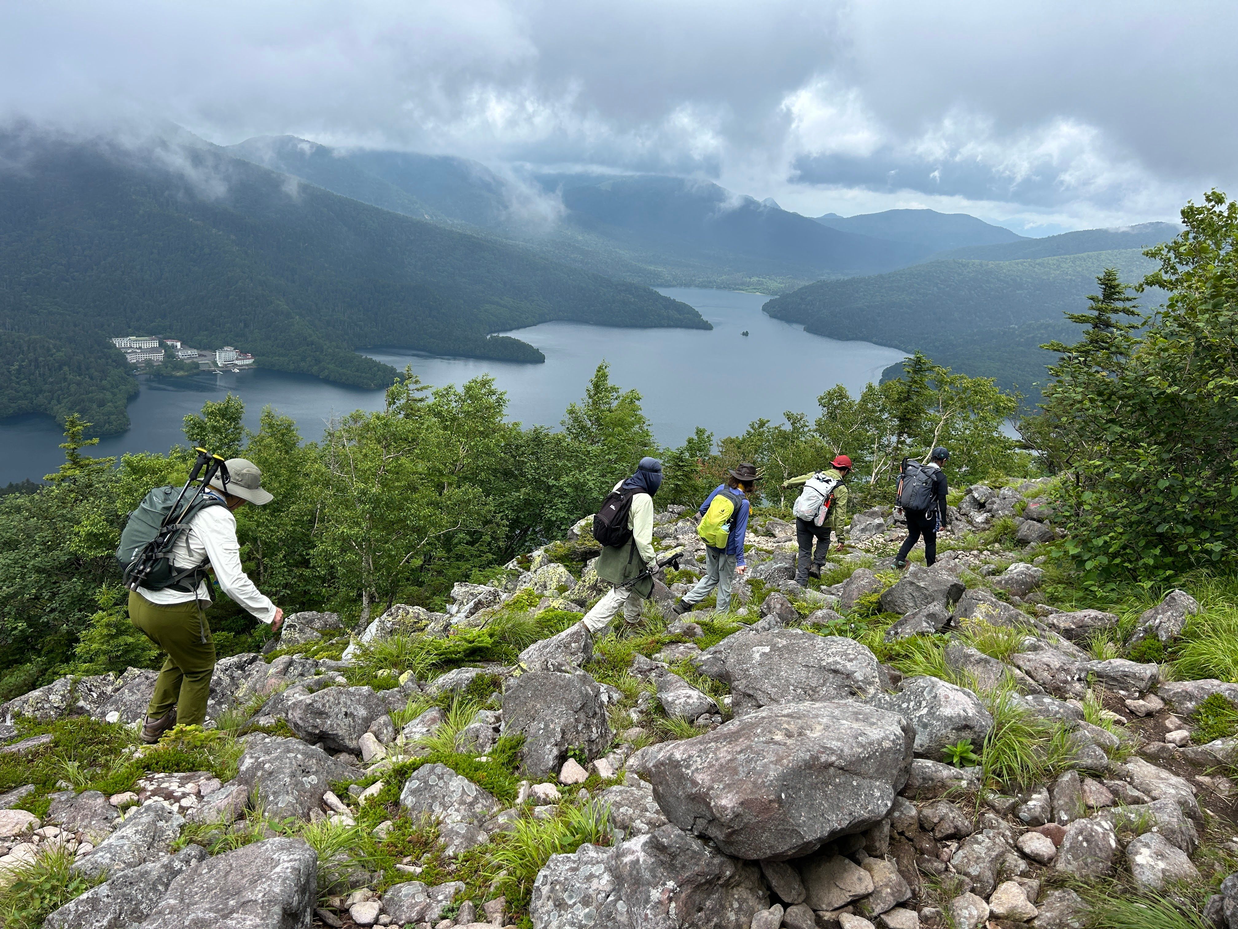 A group of hikers descend the rocky summit of Mt. Hakuunzan in Hokkaido. Below them, Lake Shikaribetsu is visible in a misty mountain valley.