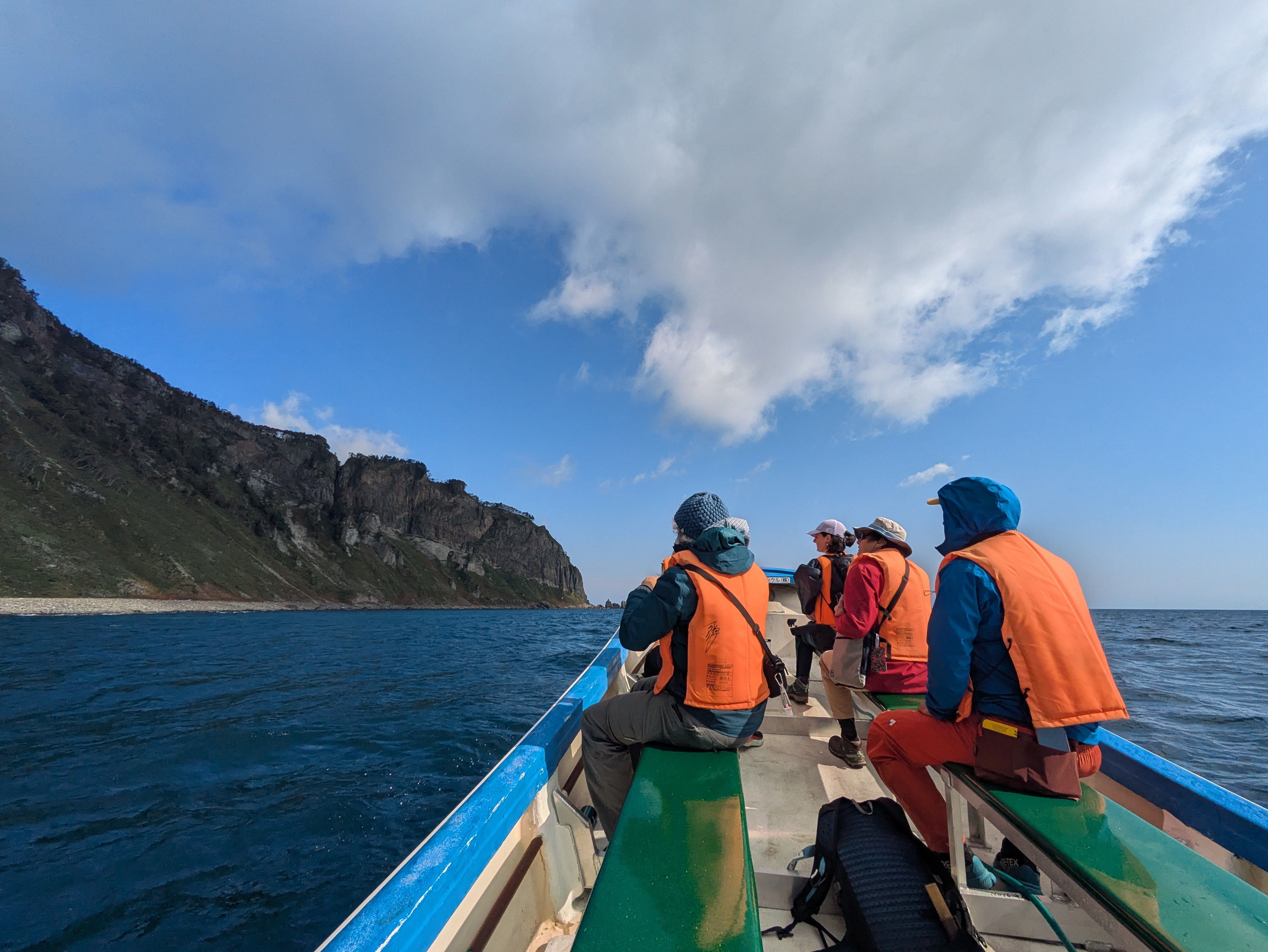 A group of people in raincoats and life jackets sit on benches in a small boat, looking out at the cliffs of the Shiretoko Peninsula in the distance. It is a very sunny day and the sea looks quite still.