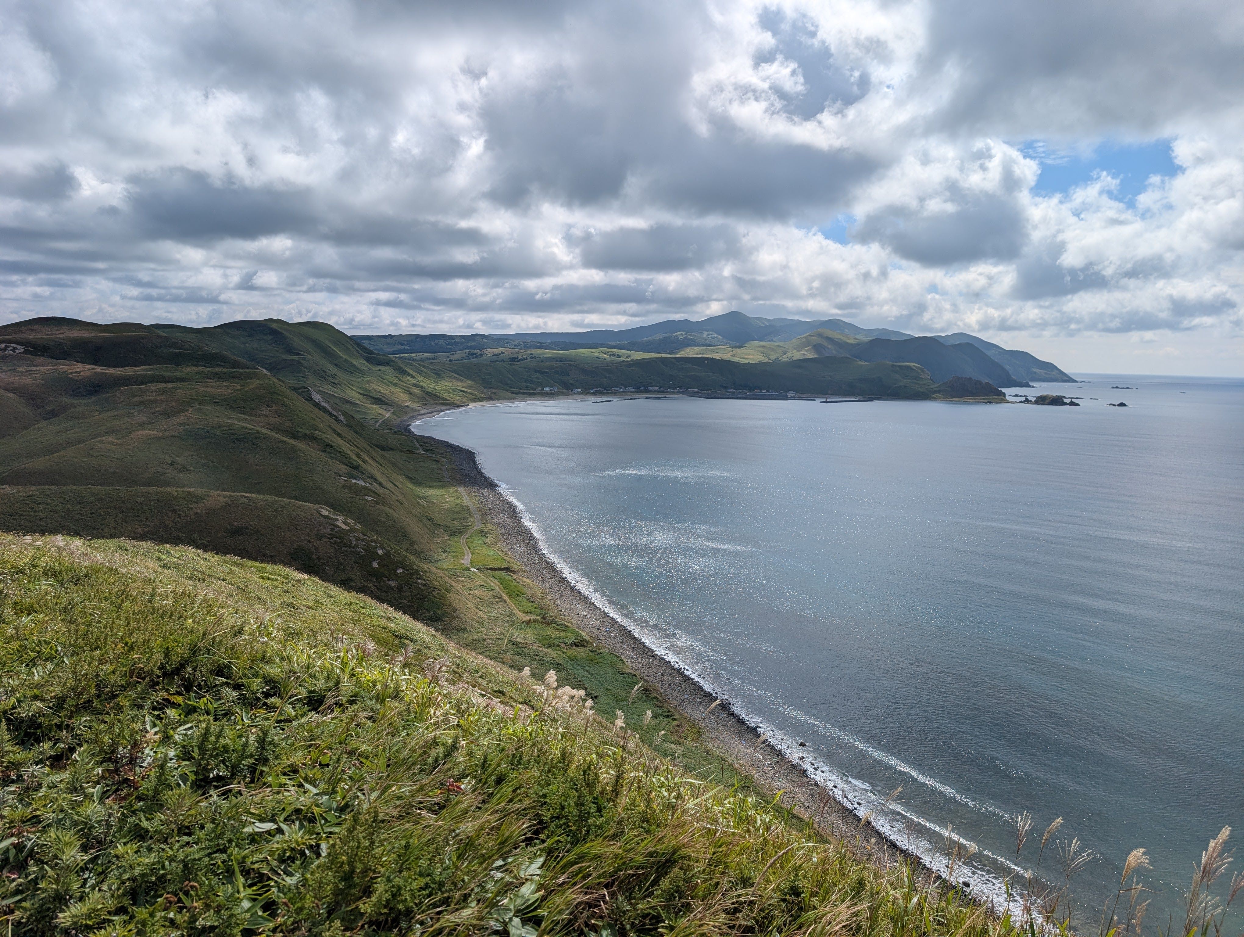 A scenic coastal view from the Misaki Meguri hiking route on Rebun Island. Lush green rolling hills slope gently down to a curved rocky beach and a vast, calm blue ocean. Distant mountains rise along the rugged coastline under a dramatic, cloud-dappled sky.