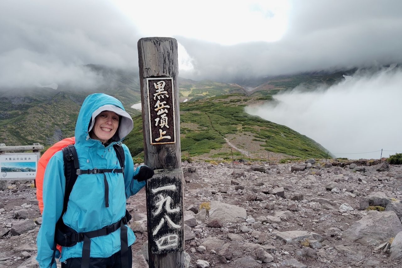 A smiling hiker in a bright blue rain jacket stands next to a wooden summit marker with Japanese characters at the top of Mt Kurodake in Daisetsuzan National Park. Behind her is a sweeping view of a rocky alpine trail, green mountain slopes, and low-hanging clouds.