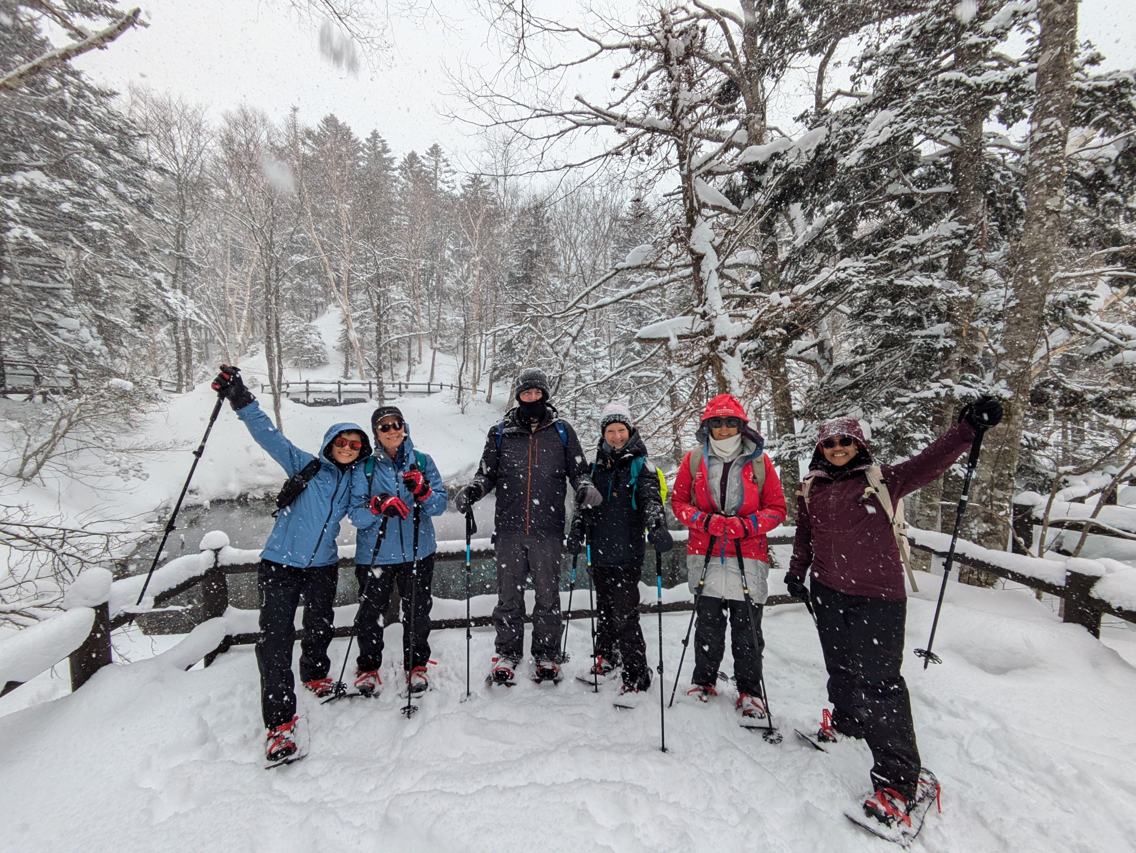 A group of snowshoers wave and smile at the camera in a snowy forest. It is snowing heavily.