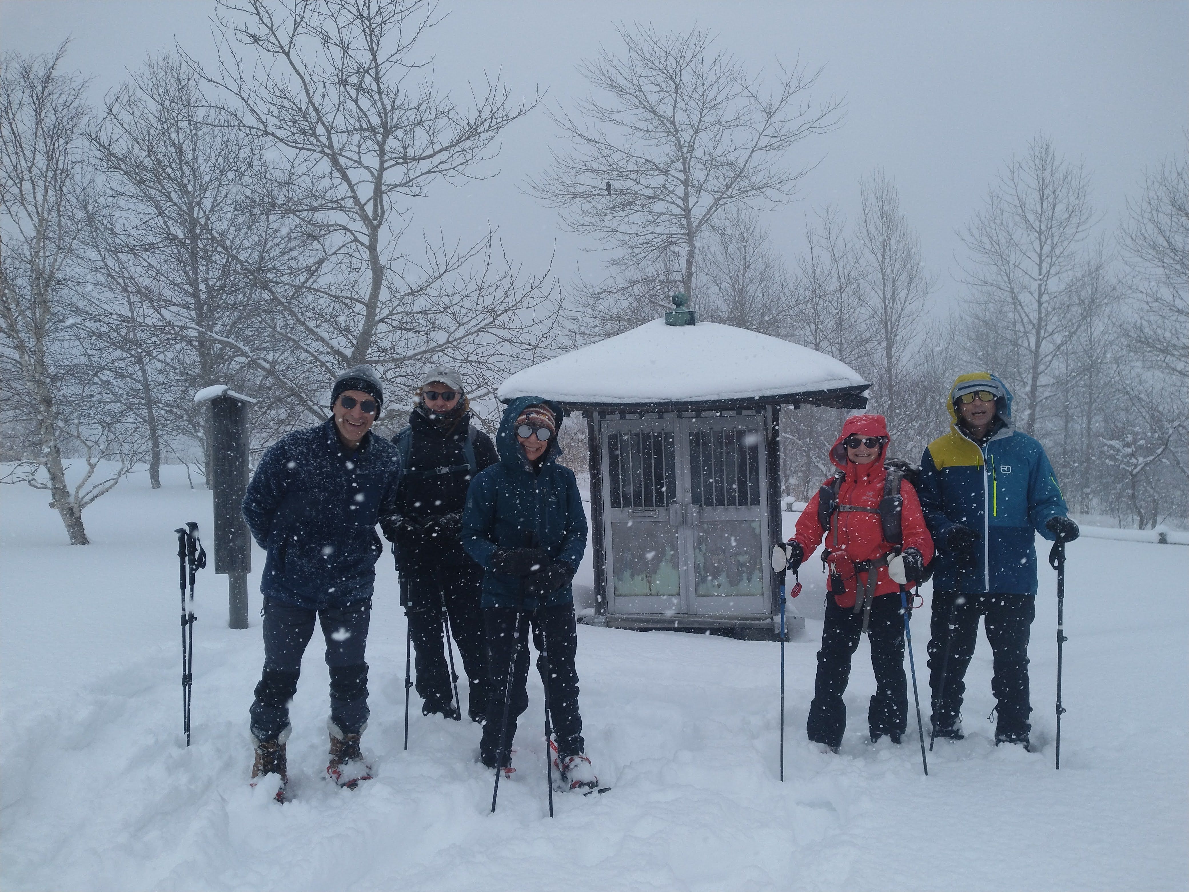A group of winter hikers smile at the camera in the snow on Mt. Usu. Snow is falling heavily around them. Behind them is a small, snow-covered temple-like building.