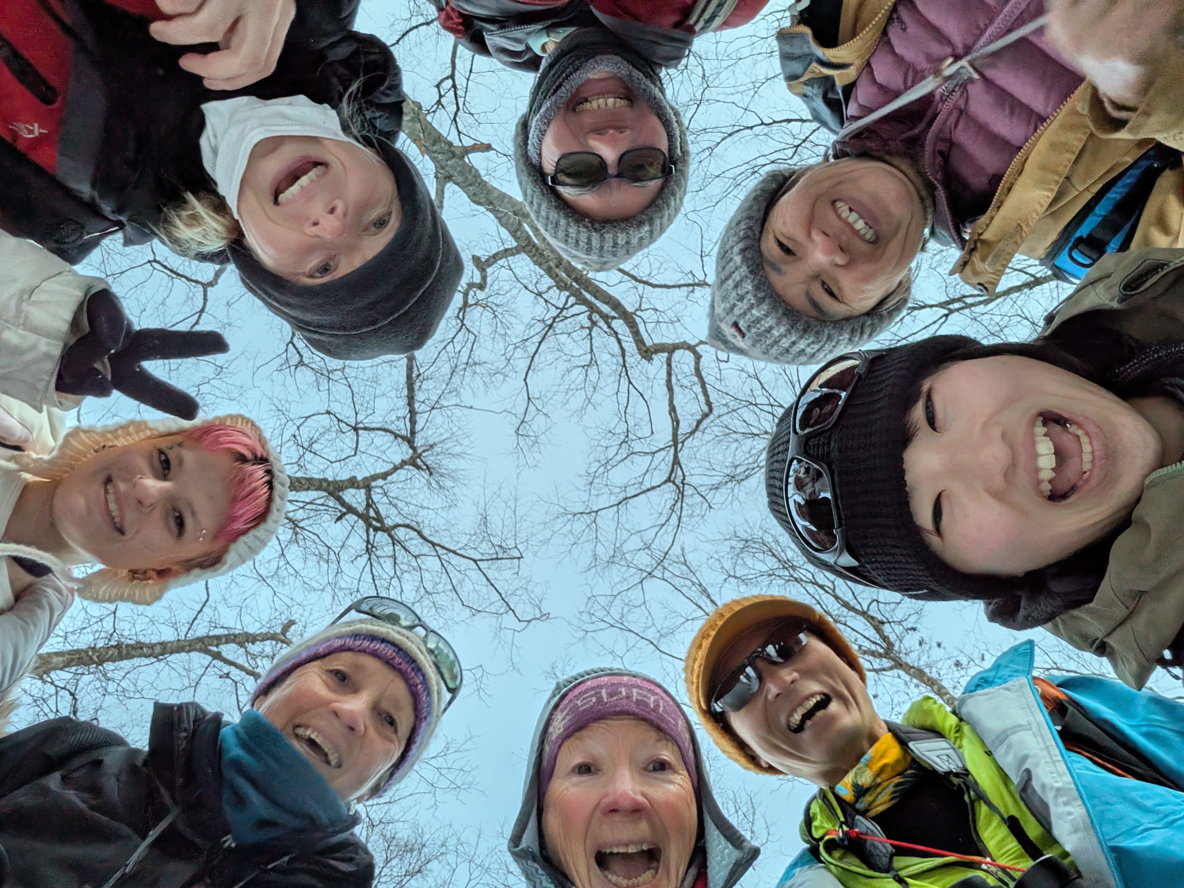 A group of people in sunglasses, woolly hats and other winter gear look down in a circle at the camera, all smiling and making faces.