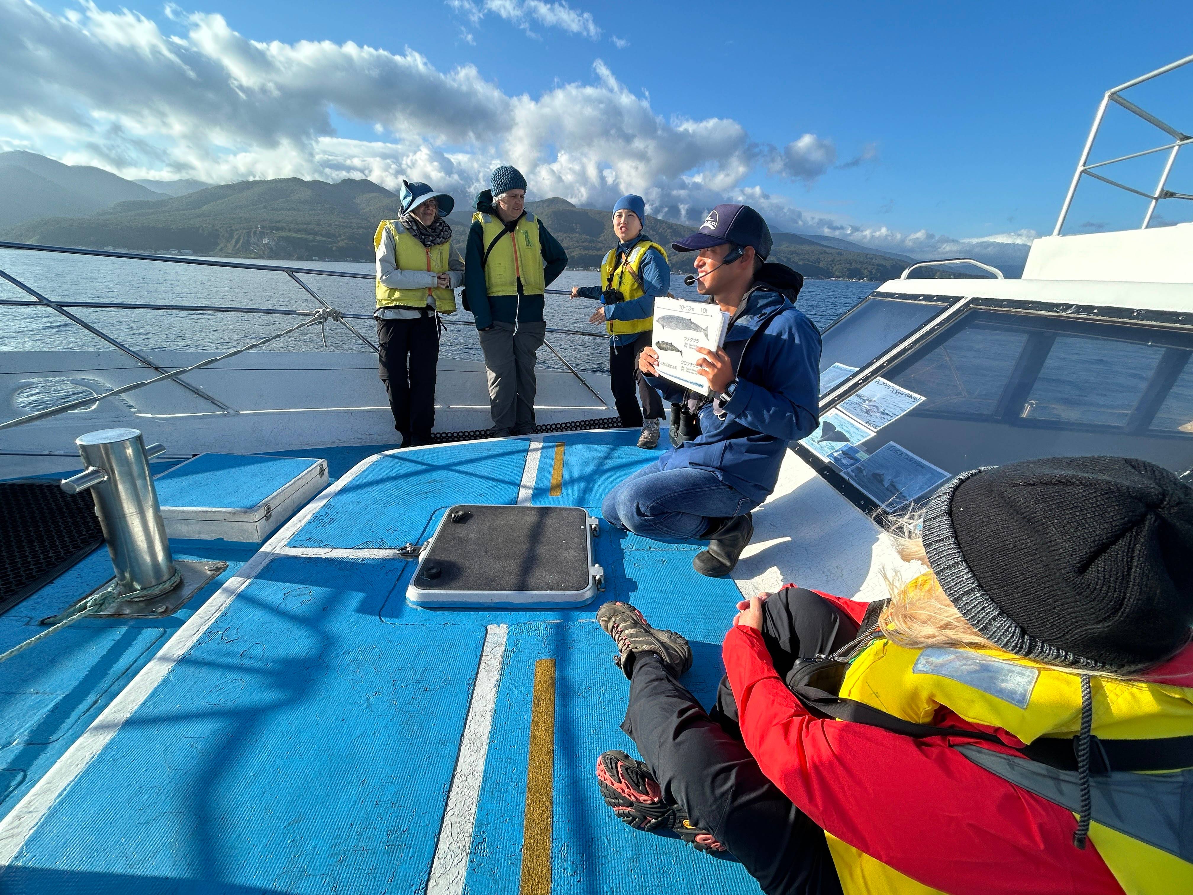 A group of guests in life jackets sit on the prow of a whale-watching cruise boat where a man holds a diagram of whales, explaining them to the guests.