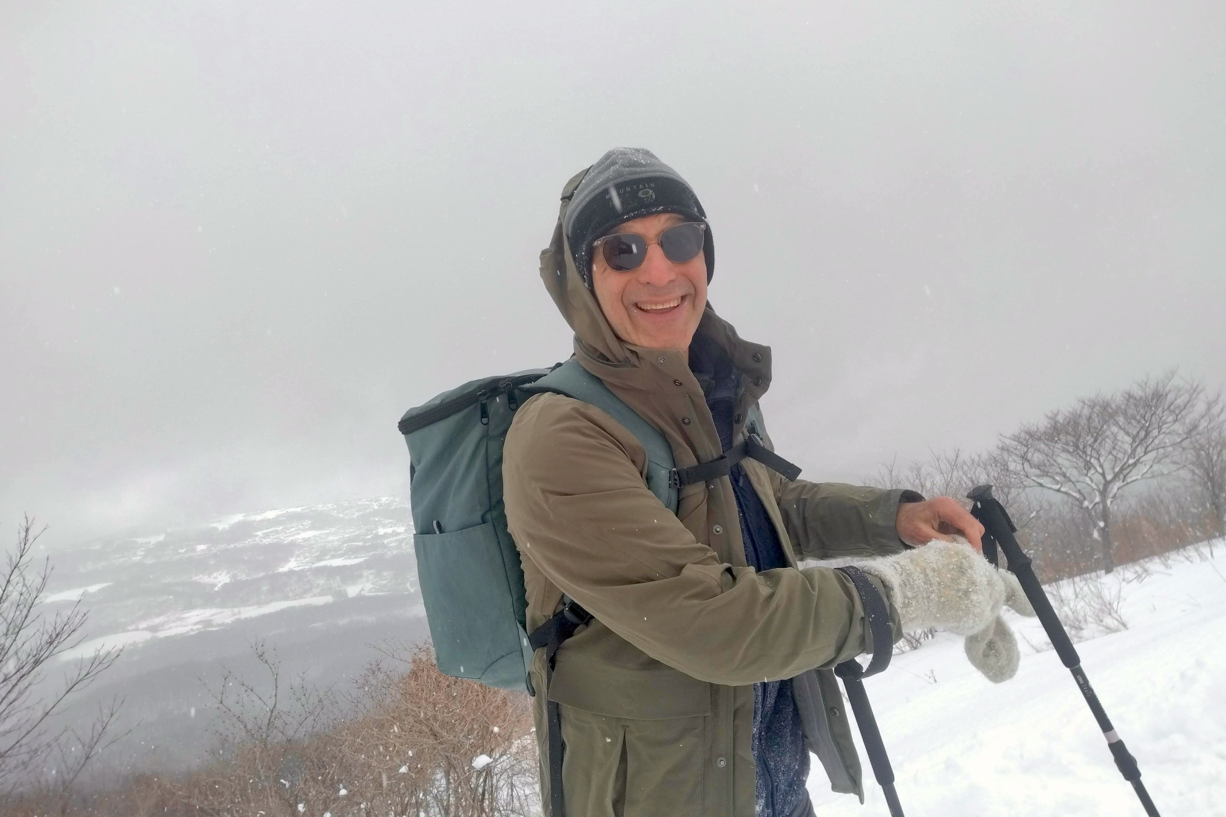 A smiling guest wearing sunglasses, a green parka, and warm mittens pauses during a winter hike at Mt. Usu. He holds trekking poles and carries a backpack, surrounded by a snowy landscape with a misty background.