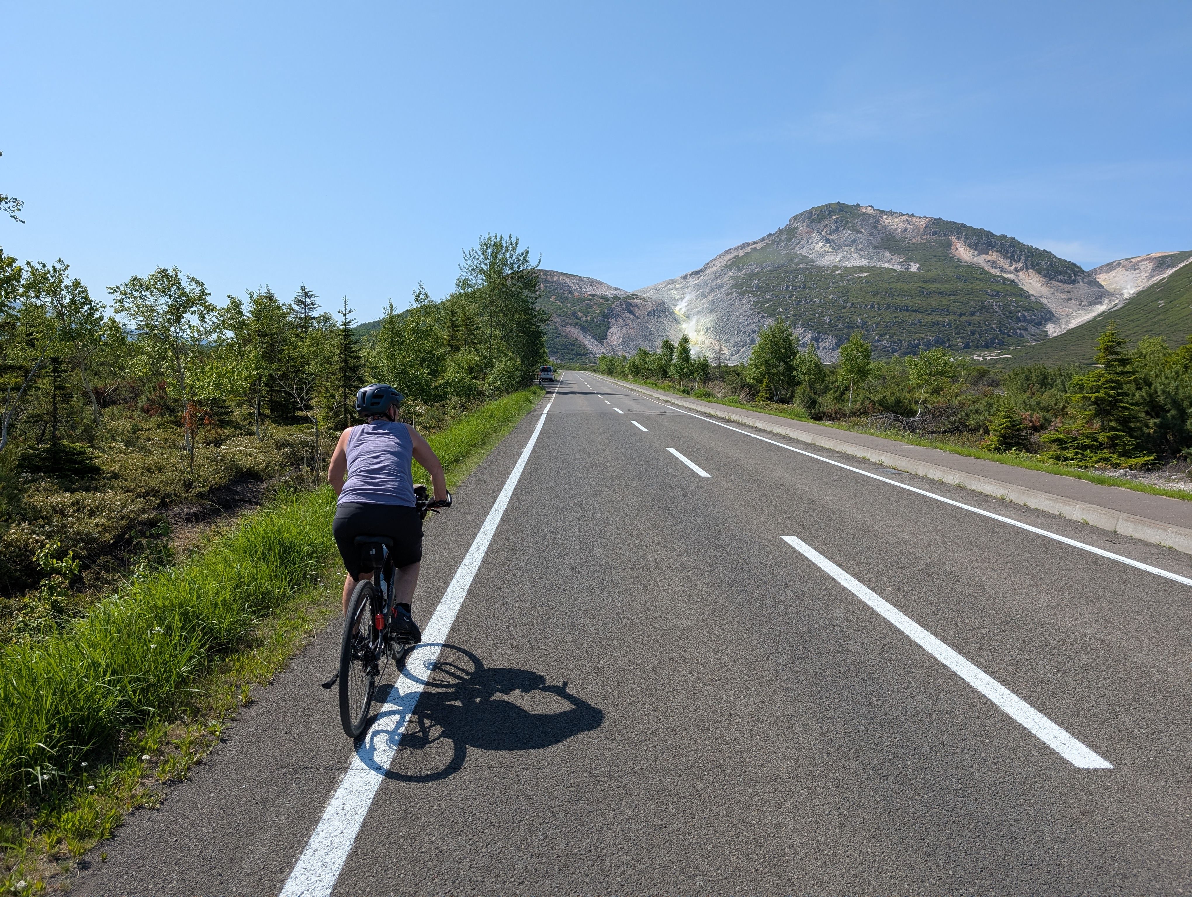 A cyclist approaches Mt. Io in east Hokkaido. It is a very sunny day and the mountain is clearly visible. Steam rises from a sulphuric vent.