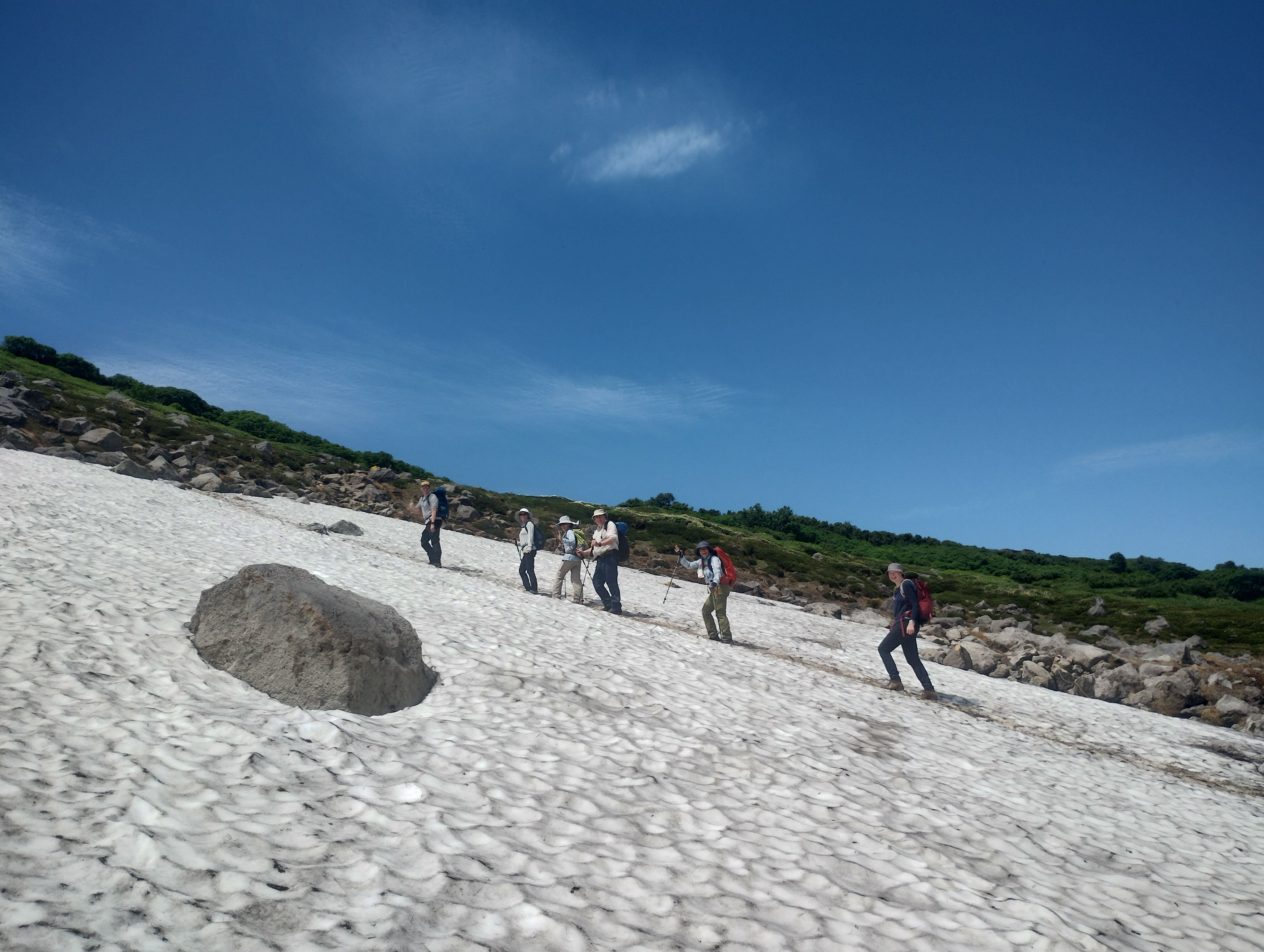 A line of hikers descends up a snowfield in Daisetsuzan National Park, Hokkaido. They are smiling and waving at the camera. It is a very sunny day.