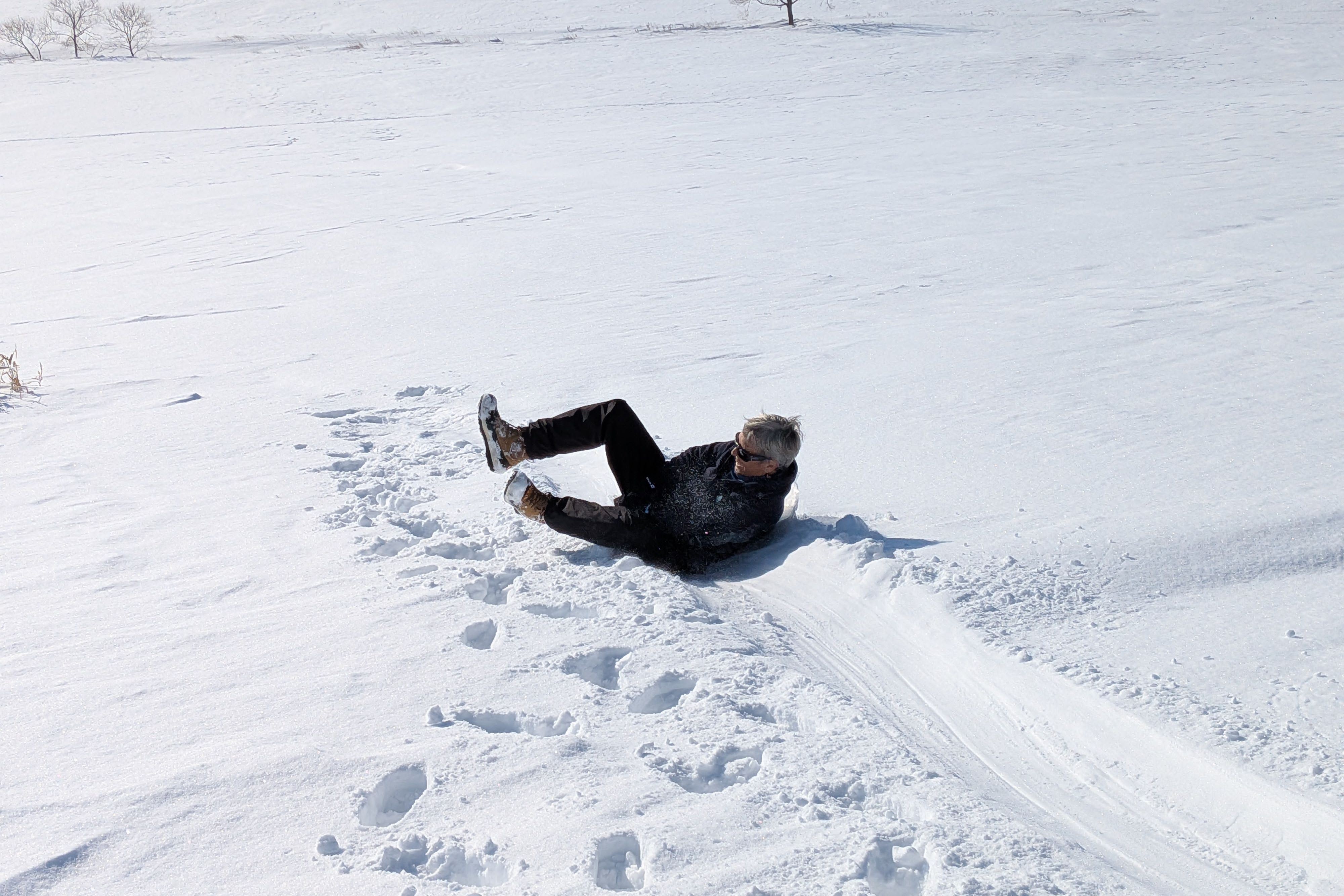 A woman sleds downhill on a snowy hill, using a small hip sled (the user sits on the sled and holds on to a handle between their legs). It looks like she is having a lot of fun.