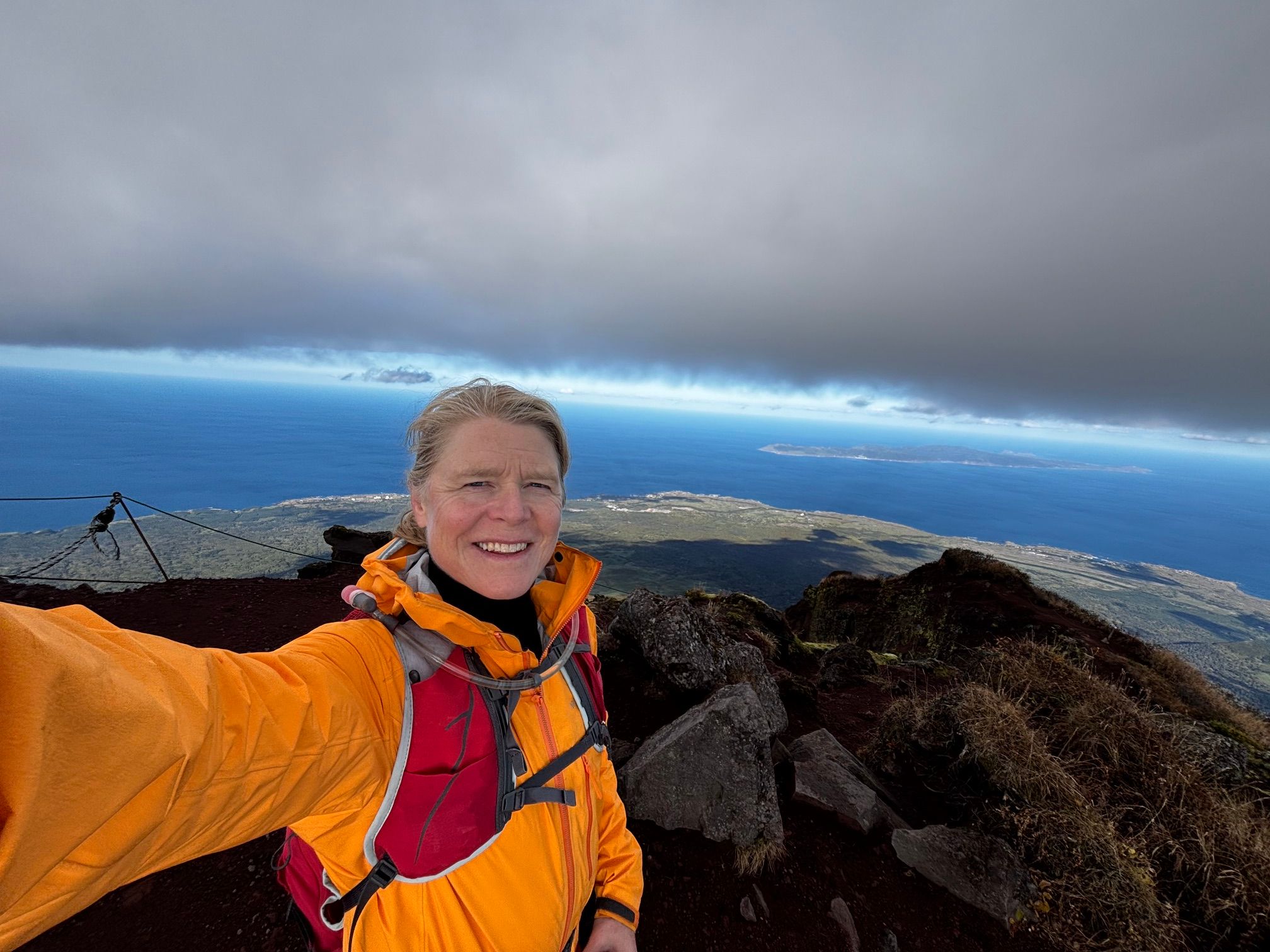 A smiling woman wearing a bright orange jacket and a red backpack takes a selfie at the rocky summit of Mount Rishiri in Hokkaido. The spectacular background reveals a sweeping panoramic view of the deep blue ocean meeting a sea of white clouds far below her.