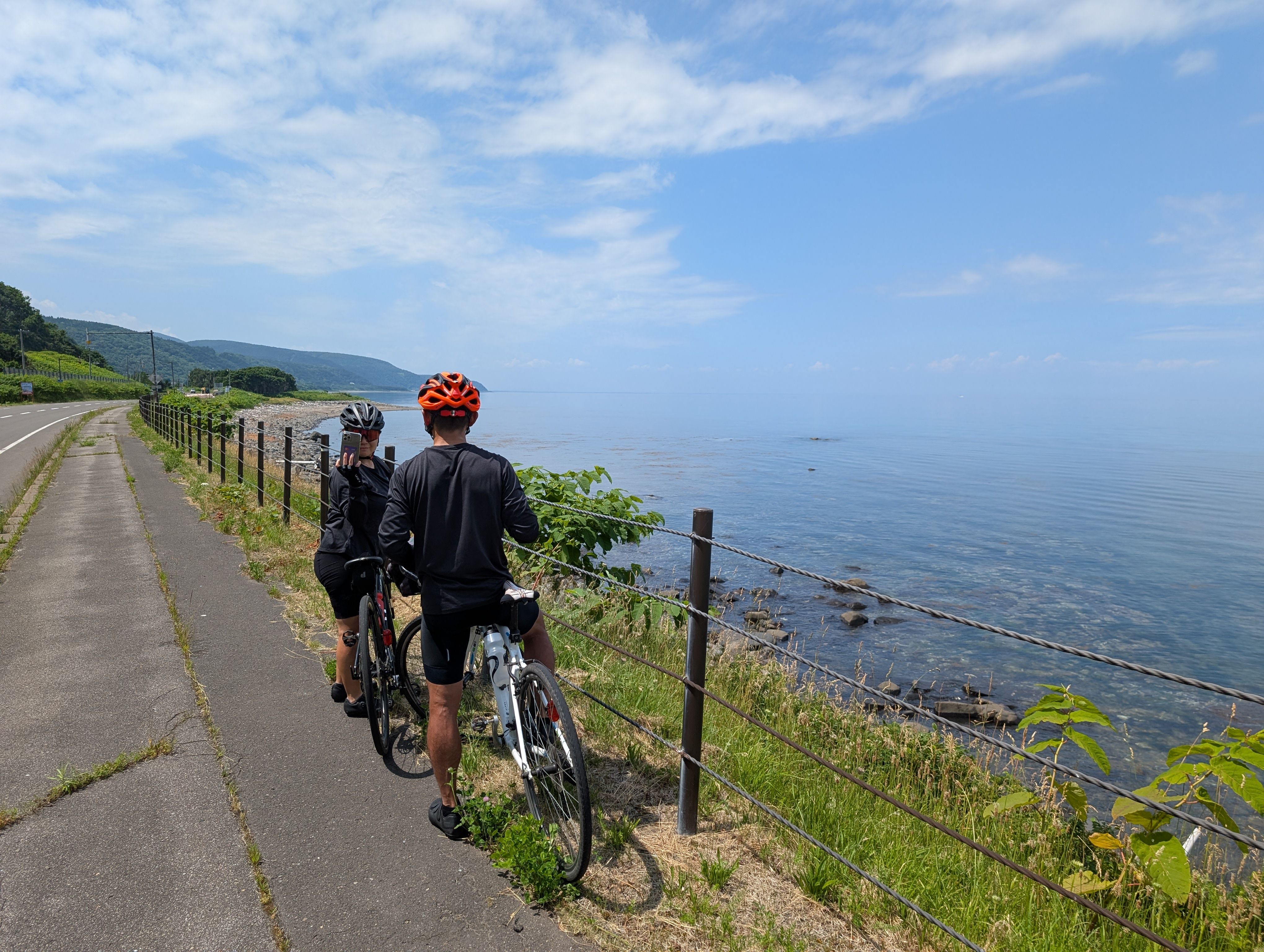 Two cyclists dismount their bikes and admire a view of the ocean. One is taking a selfie. It is a beautiful day and the sea and sky are a clear blue.