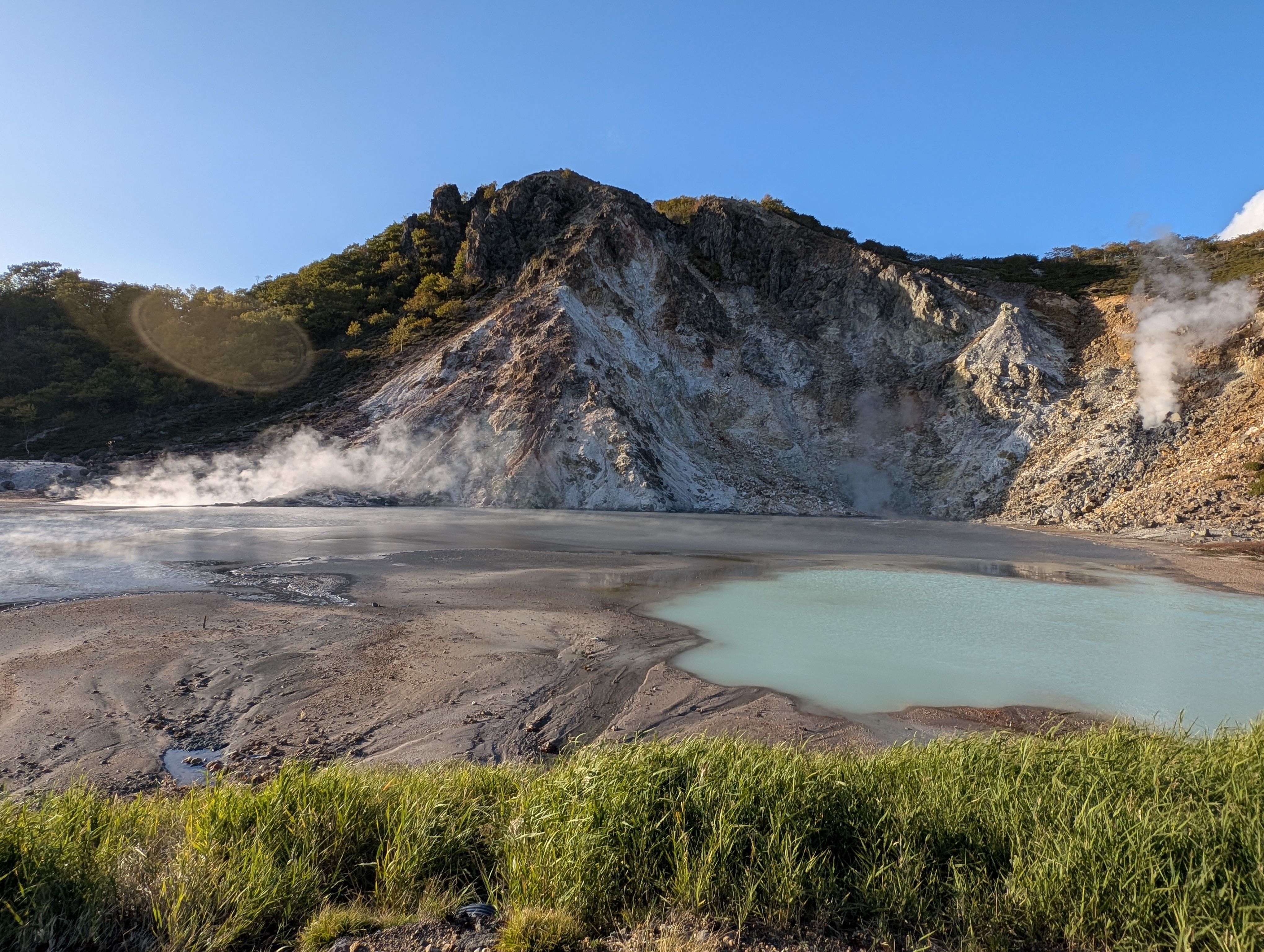 A view of a hot spring pond at Noboribetsu in Hokkaido, Japan. The hot spring pond is a bluish-green in the volcanic sand. The lip of a crater is visible in the background, steam rising from the rocks.