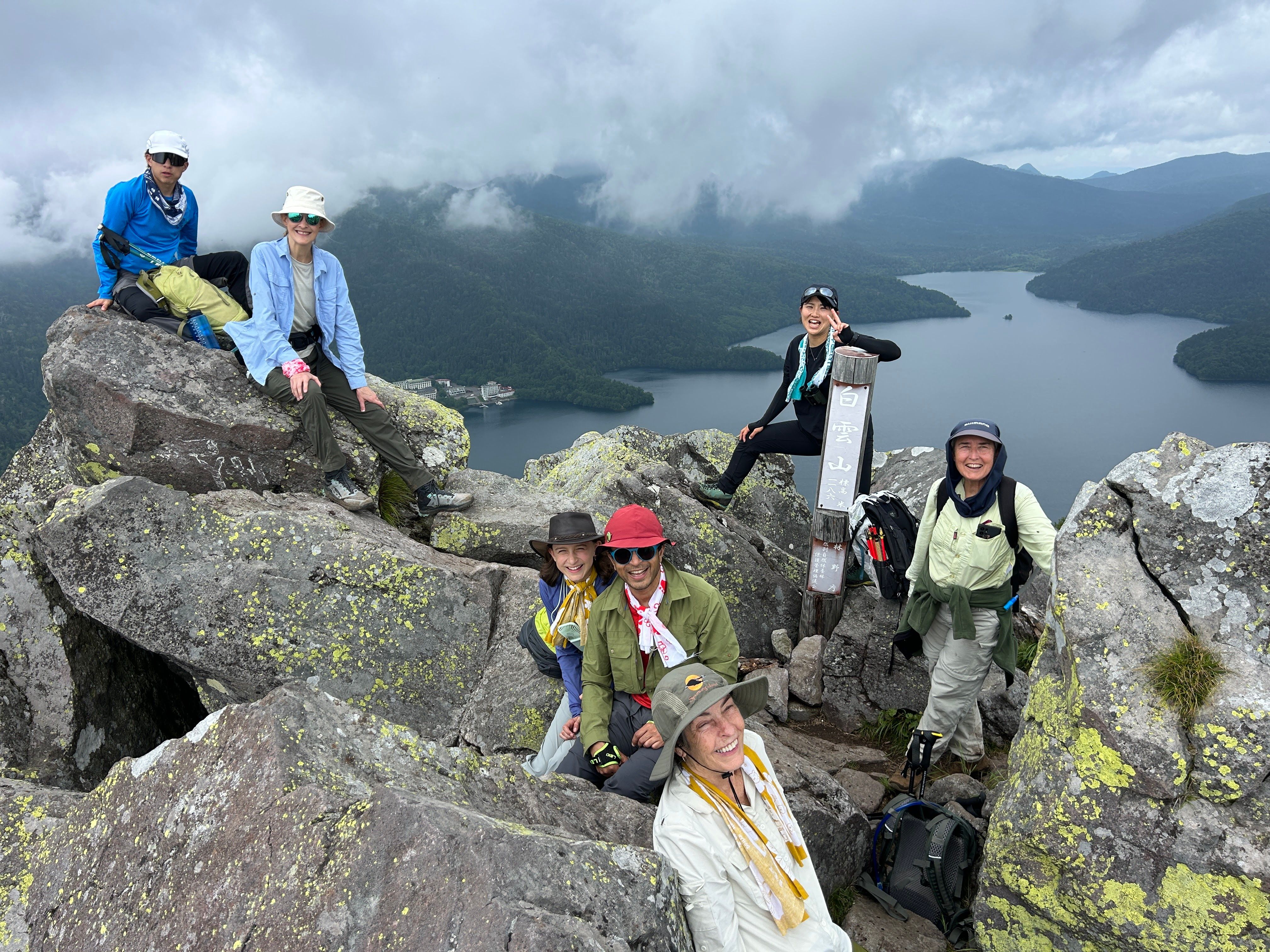 A group of hikers smile at the camera at the summit of Mt. Hakuunzan in Hokkaido. Lake Shikaribetsu is visible in the mountain valley below them, clouds hanging around the summit of the peaks beyond.