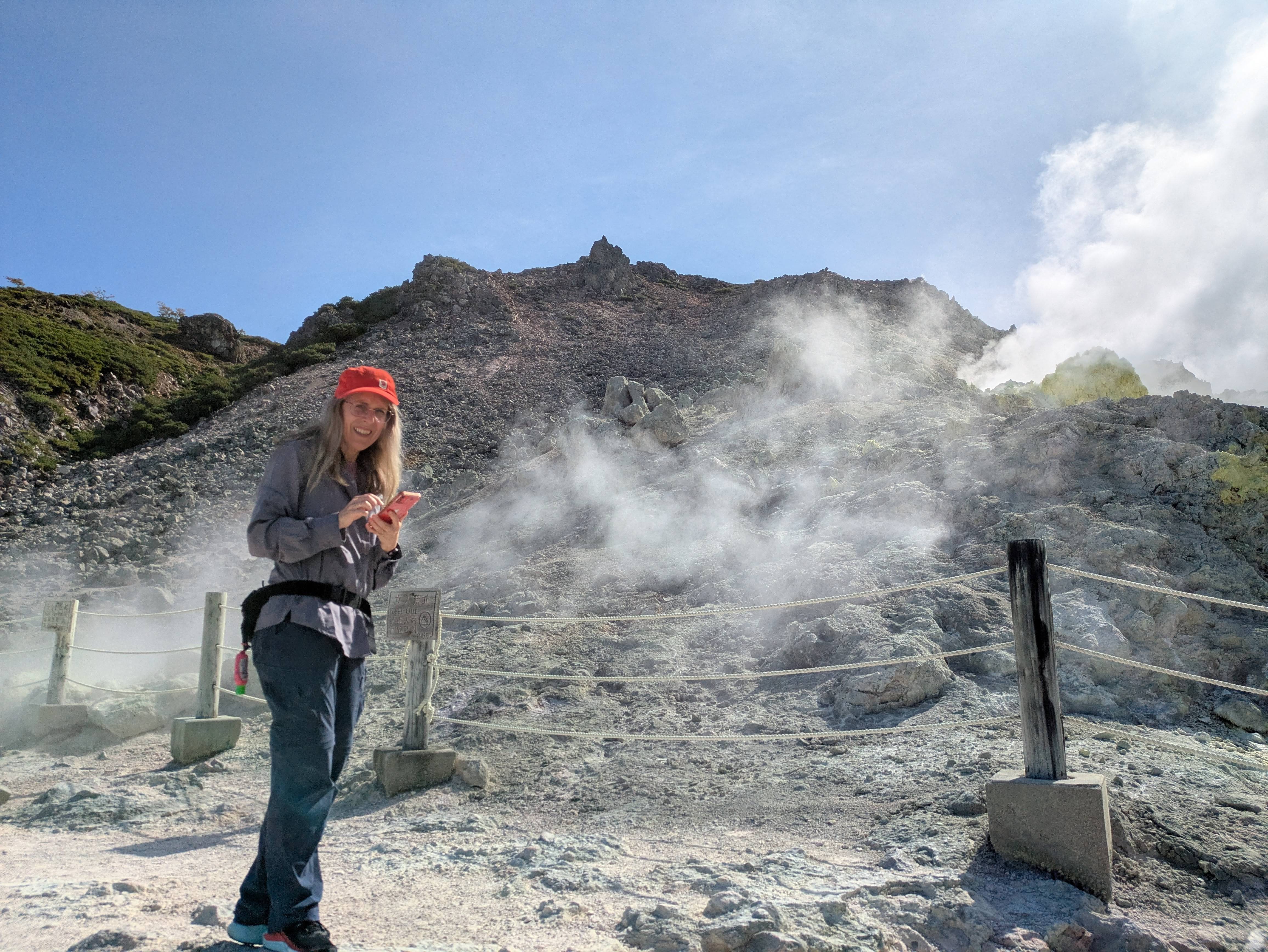 A woman smiles at the camera in front of the sulphuric vents of Mt. Io, Hokkaido. Steam rises from the rocky mountain behind her and yellow patches of sulphur mineral deposits are visible.