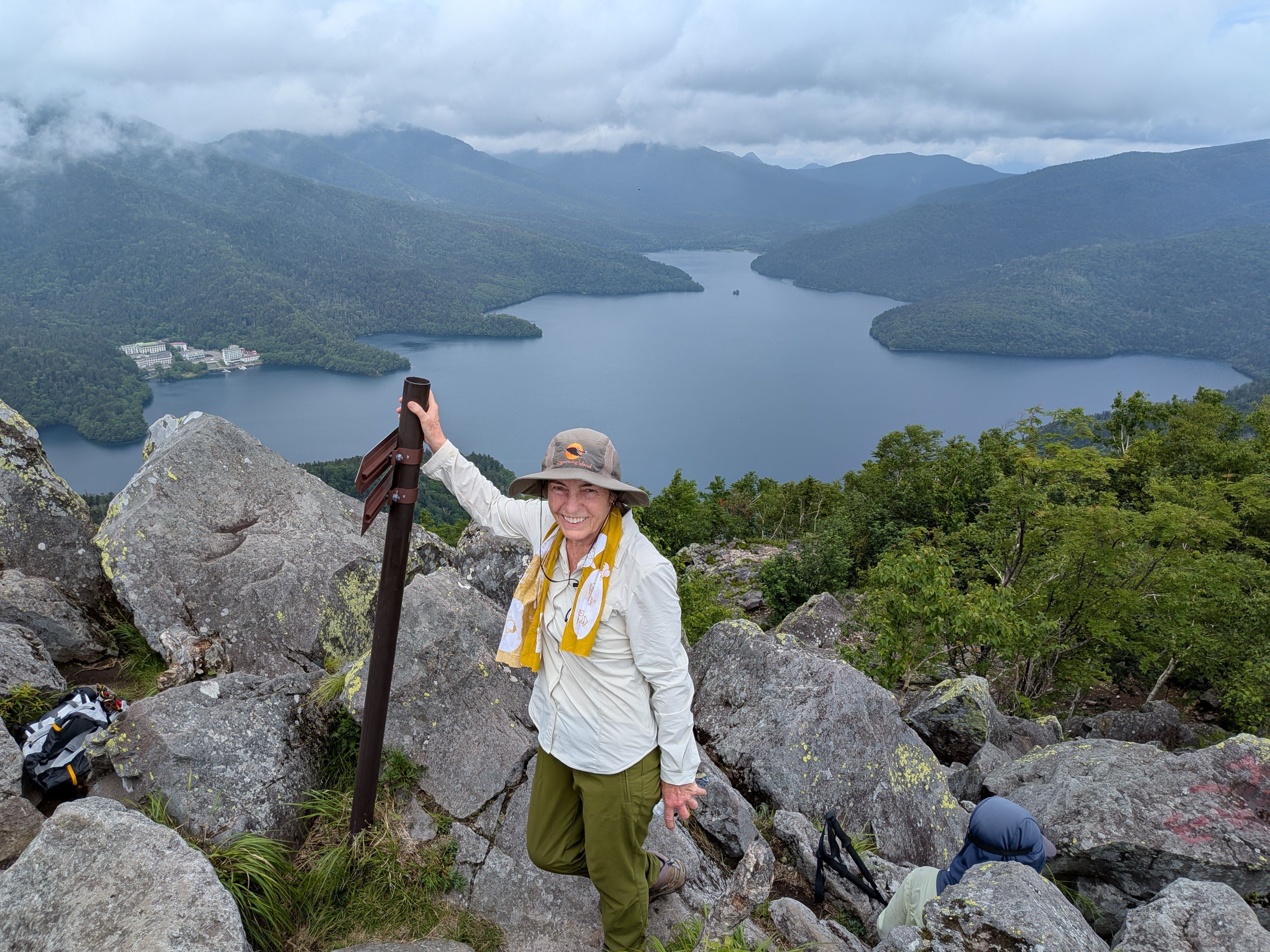 A smiling female guest stands on the rocky summit of Mt Hakuunzan, holding a trail signpost. Behind her is a breathtaking panoramic view of Lake Shikaribetsu and lush forested mountains under a cloudy sky.