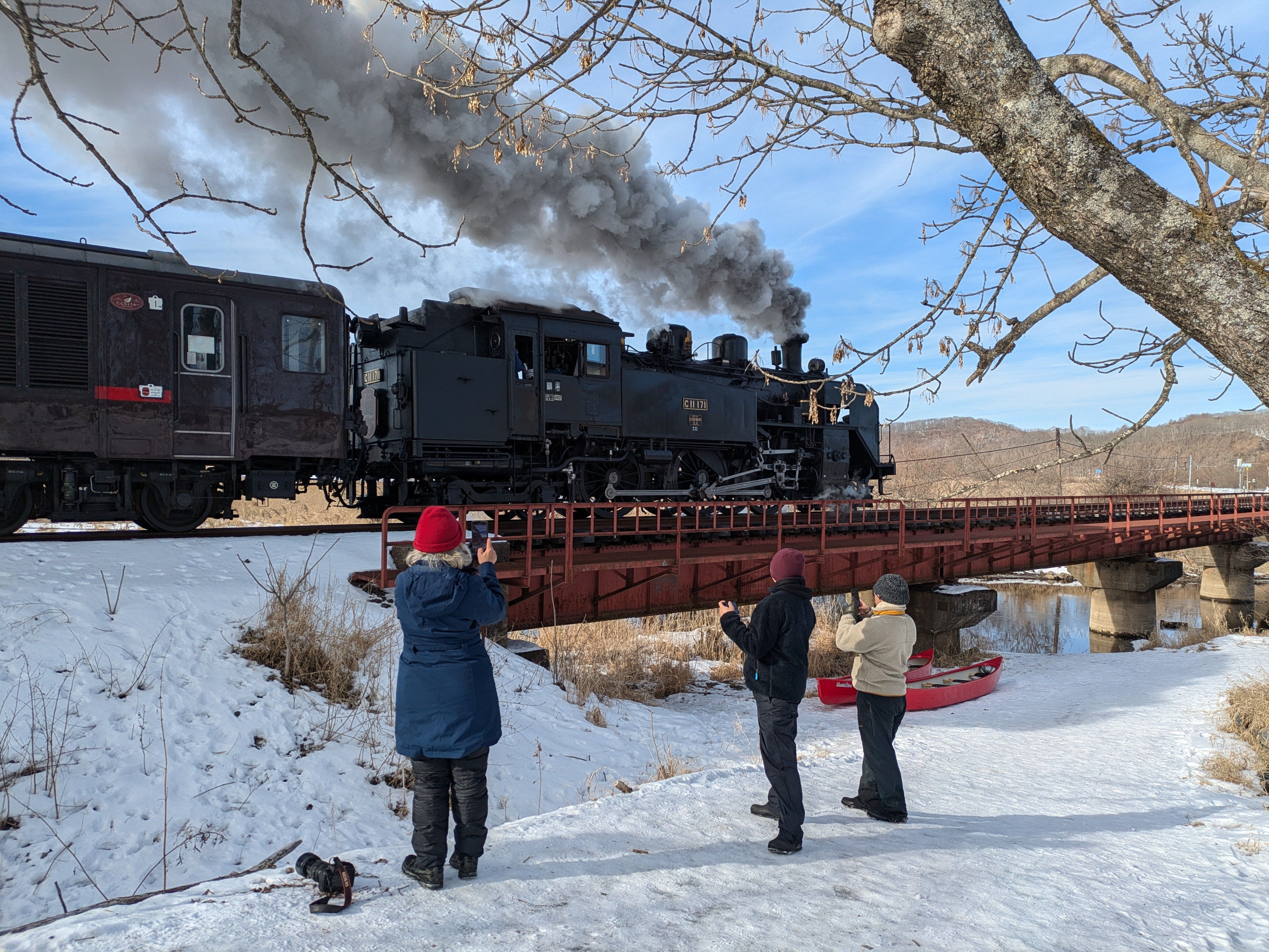 Three people take photographs of a steam train as it crosses a bridge in front of them. It is midwinter and there is snow on the ground.