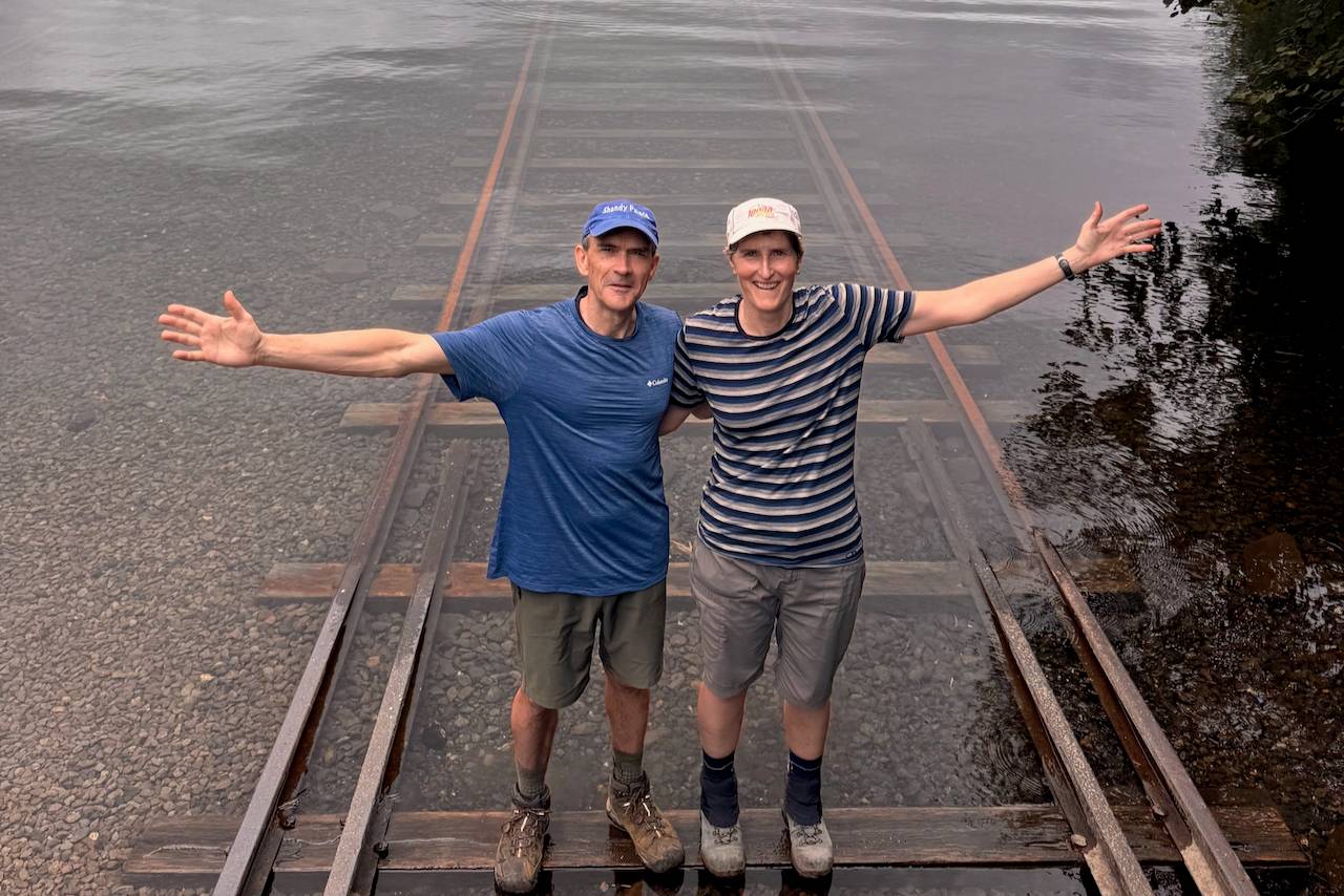 A couple, Alison and Mike from New Zealand, stand on a submerged railway track in the clear water of Lake Shikaribetsu, Hokkaido. They are smiling broadly and have their arms outstretched. They are both wearing hats, t-shirts, and shorts.