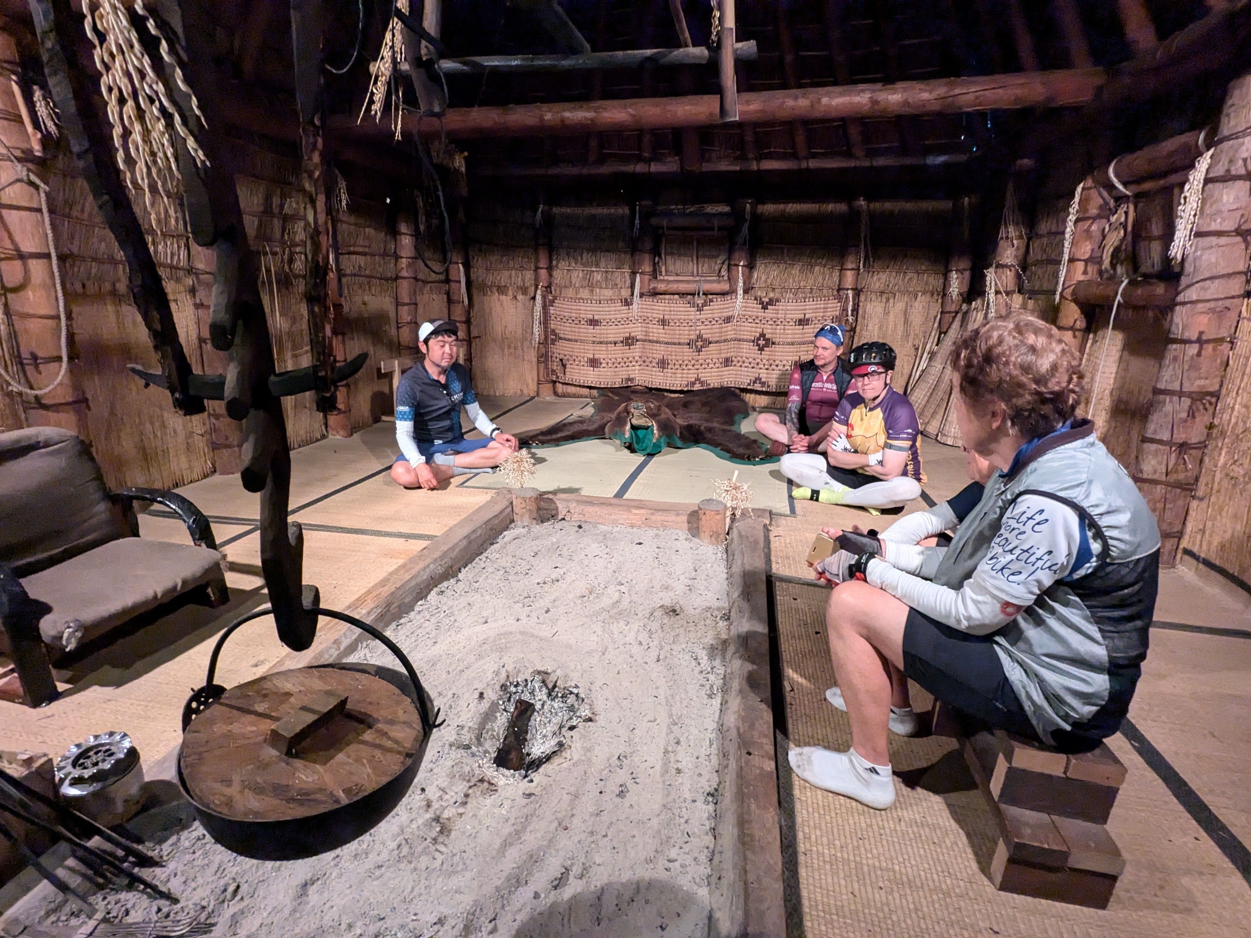 A group of people sit inside a cise, a traditional Ainu dwelling made from reeds and bamboo leaves. An ashy hearth is in the middle of the room with a pot suspended over it.