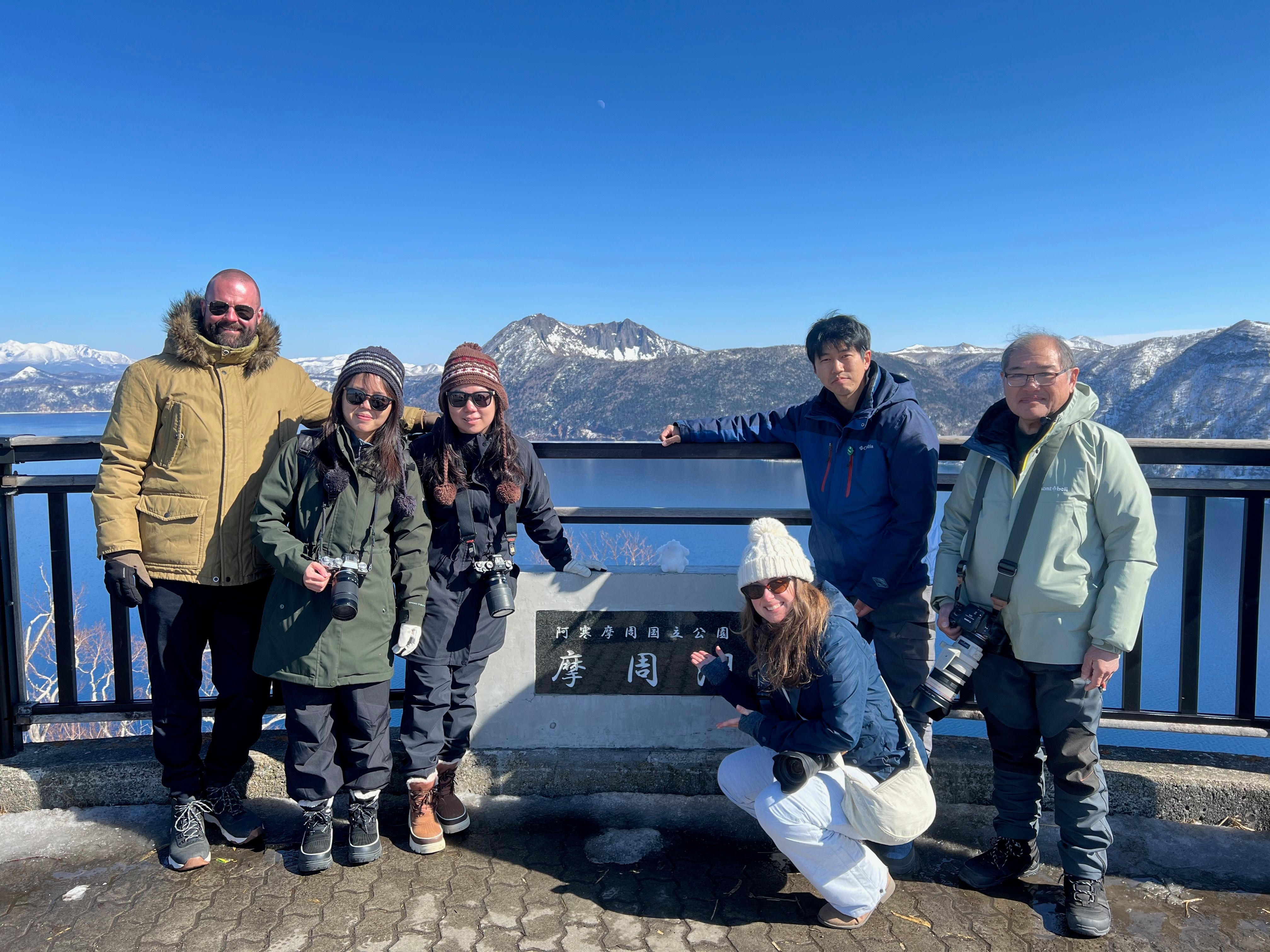 A group of people smile at the camera and crouch or stand around a sign overlooking Lake Mashu in Hokkaido. The sign, in Japanese, reads "Akan-Mashu National Park: Lake Mashu".