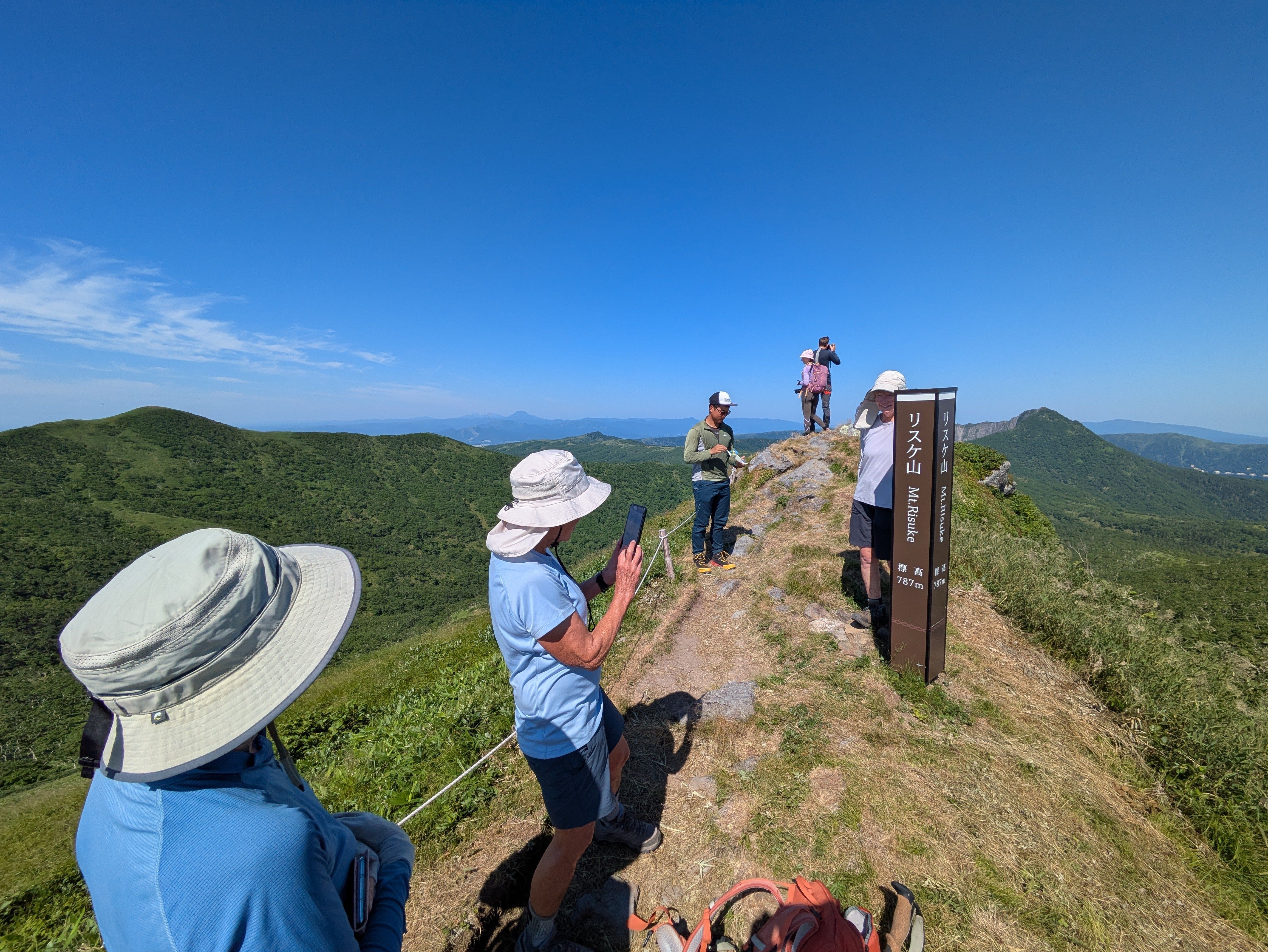 A group of hikers stand at the summit of Mt. Risuke, taking photographs of the view. It's a very sunny day and other mountains are clearly visible in the distance.