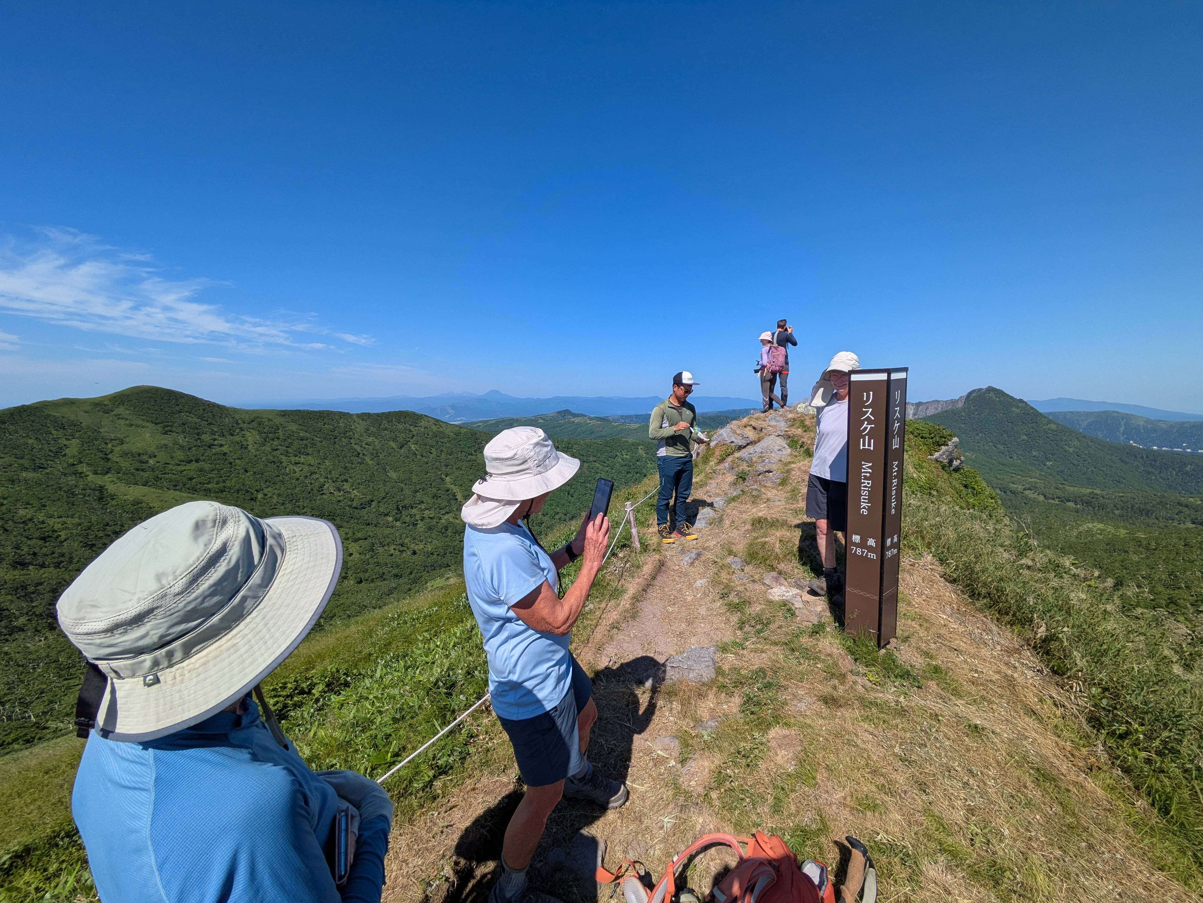 A group of hikers stand at the summit of Mt. Risuke, taking photographs of the view. It's a very sunny day and other mountains are clearly visible in the distance.